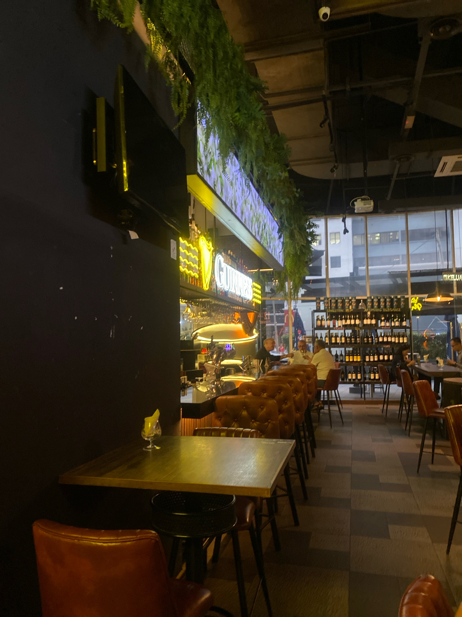 The image depicts the interior of a bar or restaurant, likely located in Kuala Lumpur, Malaysia, given the context. The scene is dimly lit, with warm lighting emanating from neon signs and ambient lamps. In the foreground, a brown leather armchair is visible, situated at a wooden table with a small glass containing a yellow garnish. Behind this, a row of similar leather chairs faces a bar area.
The bar itself features a prominent neon Guinness sign, with bottles and glassware arranged behind it. Further back, a large window looks out onto a city street at night, with buildings and streetlights visible. A wall on the left is painted dark, with a television mounted on it. The ceiling is industrial, with exposed beams and lighting fixtures, including a projector. A prominent feature of the decor is hanging greenery, adding a touch of nature to the urban setting. Several patrons are seated at tables and the bar, some engaged in conversation. Bottles of wine are displayed on shelves in the background. The overall atmosphere suggests a sophisticated yet relaxed establishment.