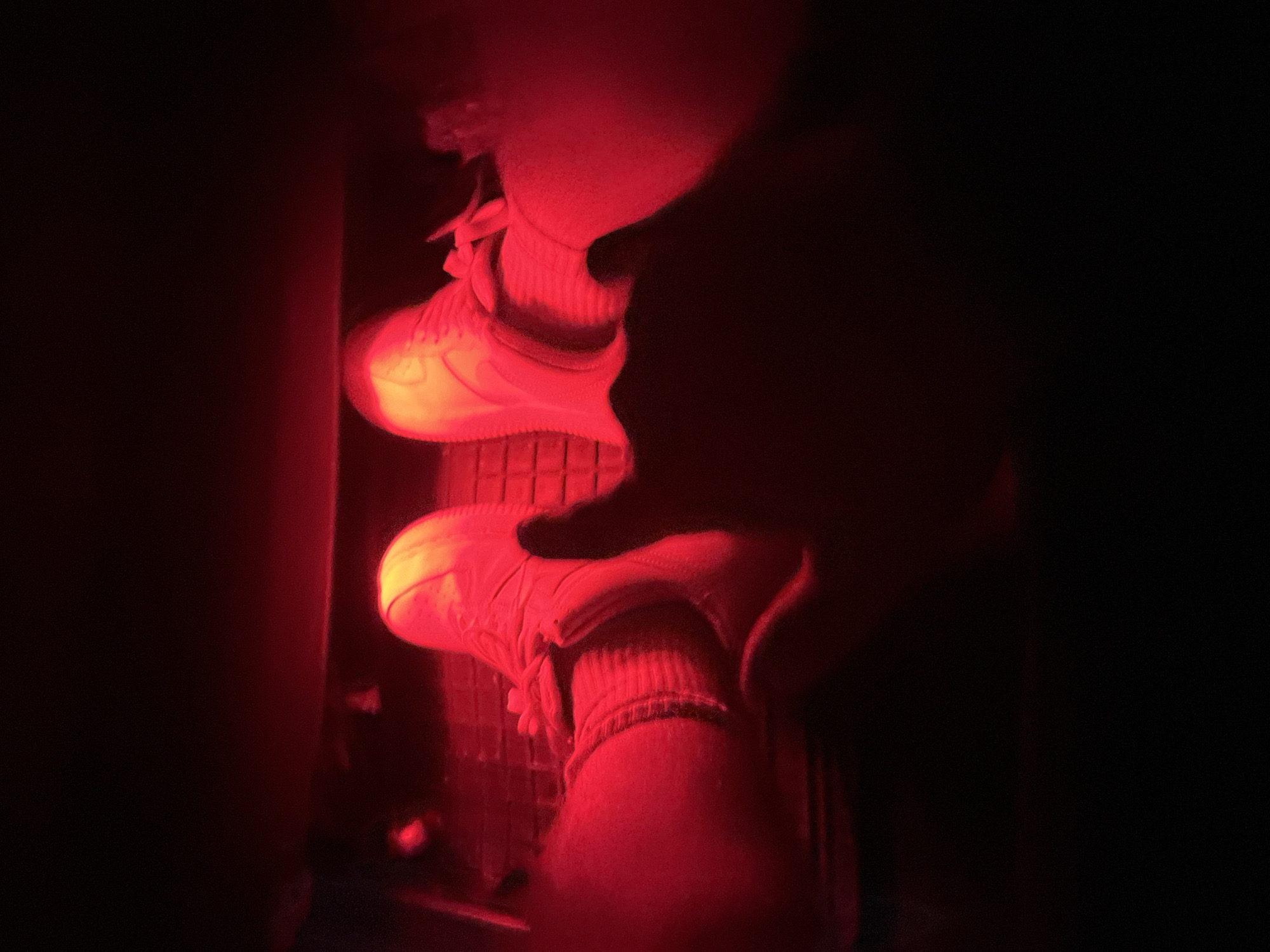 The image shows a close-up view of a person's feet in sneakers, illuminated by red light. The feet are positioned as if resting on the floor pedals of a vehicle. The left foot is slightly angled towards the viewer, while the right foot is more directly positioned. A hand is also visible in the foreground, partially obscuring the right foot, with the thumb extended upwards. The overall scene is dark, with the red light casting strong shadows and highlighting the textures of the shoes and socks. There are no discernible features of the city of Zlatograd, Bulgaria, visible in this image.