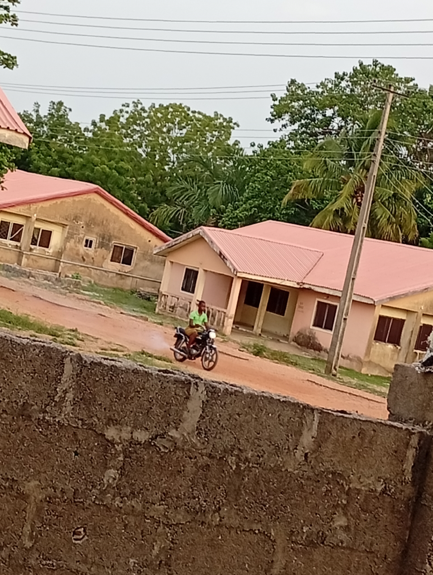 A person on a motorcycle is moving on a dirt road between two single-story buildings with red roofs in a location identified as Unknown, Unknown. The buildings have visible windows and neutral-colored exterior walls. Overhead, electrical wires are strung between utility poles, and trees populate the background. A section of a weathered concrete wall is in the foreground.