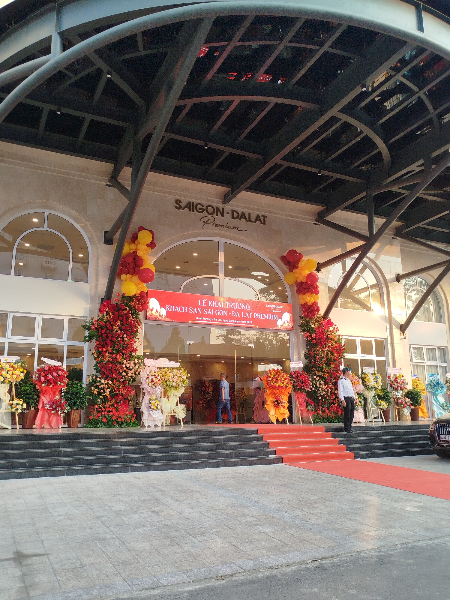 The image shows the entrance of the Saigon-Dalat Premium hotel in Da Lat, Vietnam. The hotel's name is displayed prominently above the entrance. Red and yellow balloons and flower arrangements flank the entrance. A red carpet extends up the stairs leading into the hotel. People are visible near the entrance. A sign hanging above the entrance reads "LỄ KHAI TRƯƠNG KHÁCH SẠN SÀI GÒN - ĐÀ LẠT PREMIUM, Xuân Hương - Đà Lạt, ngày 02 tháng 4 năm 2026". A vehicle partially visible on the right side of the image shows a license plate of "49A-11". The building has an arched roof and large glass windows. The sky is overcast.