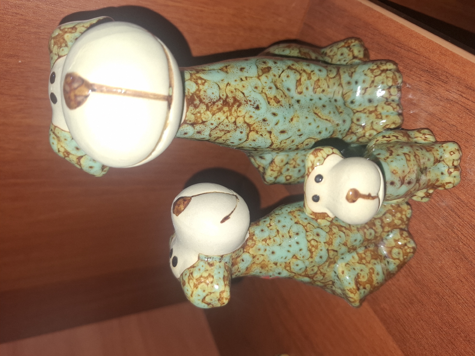 Three ceramic dog figurines are displayed on a wooden shelf. The largest figurine stands upright, with a wide, white face featuring black dots for eyes and a brown, vertical marking for a nose. Its body is teal with brown speckles. To its left is a medium-sized figurine, also teal with brown speckles, facing to the right with a white head and similar facial features to the largest one. In front of these two is a smaller figurine, resembling the others in style, with its white head facing forward and black dot eyes. The background is a dark red, wooden surface, likely a wall or shelf. The scene is set indoors on a wooden shelf. There is no indication of the city of Almaty, Kazakhstan.