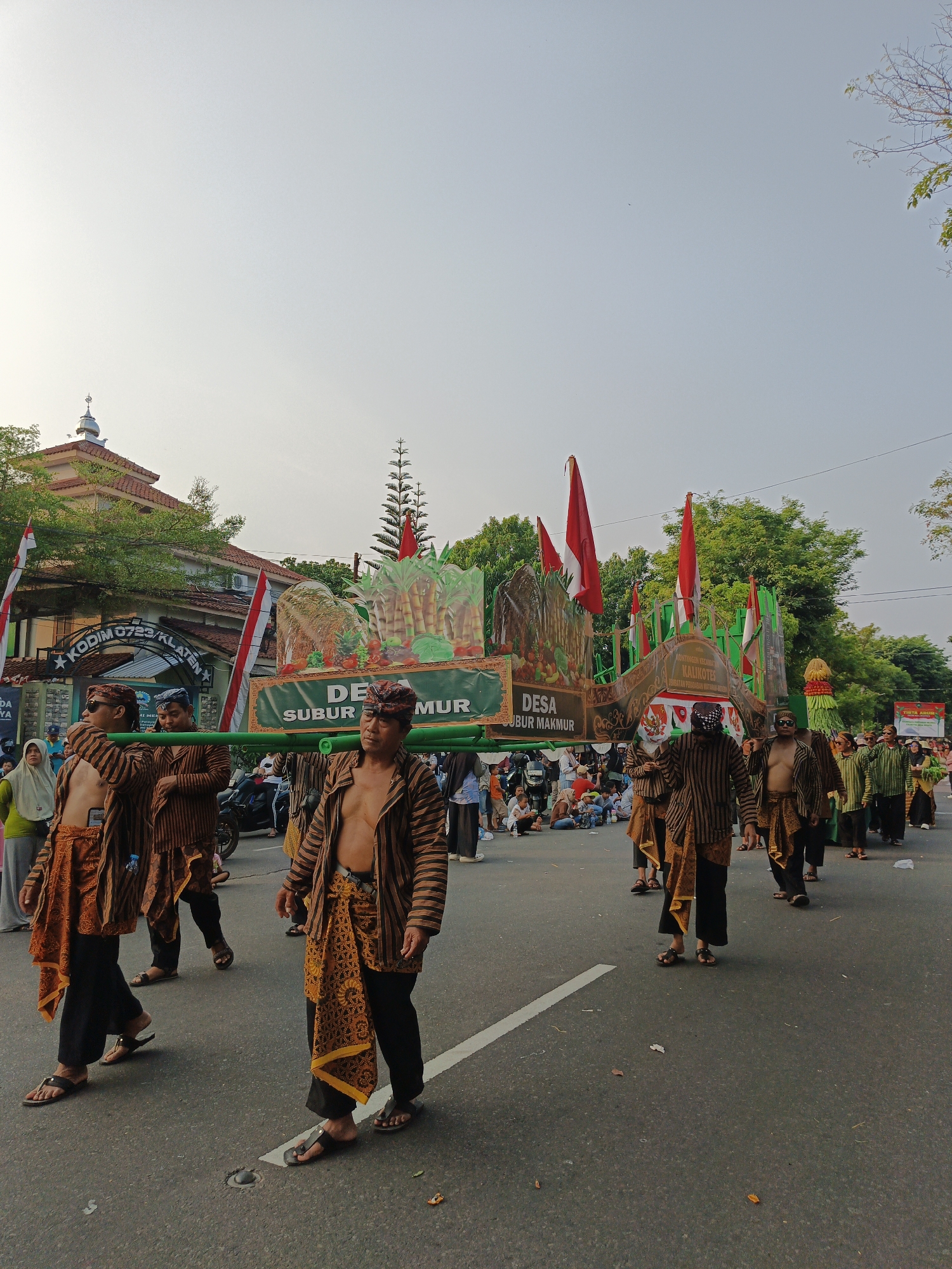 A procession is taking place on a paved road in Klaten, Indonesia, likely in front of a building identified by a sign as "KODIM 0723/KLATEN". Several men, dressed in traditional Javanese clothing consisting of striped jackets and patterned sarongs, are carrying a large, elaborately decorated float. The float features depictions of sugarcane, fruits, and possibly a landscape, adorned with red and white Indonesian flags. Spectators, some seated and others standing, line the side of the road, observing the parade. A building with a dome, resembling a mosque, is visible in the background, along with trees and the sky. The overall scene suggests a cultural event or festival.