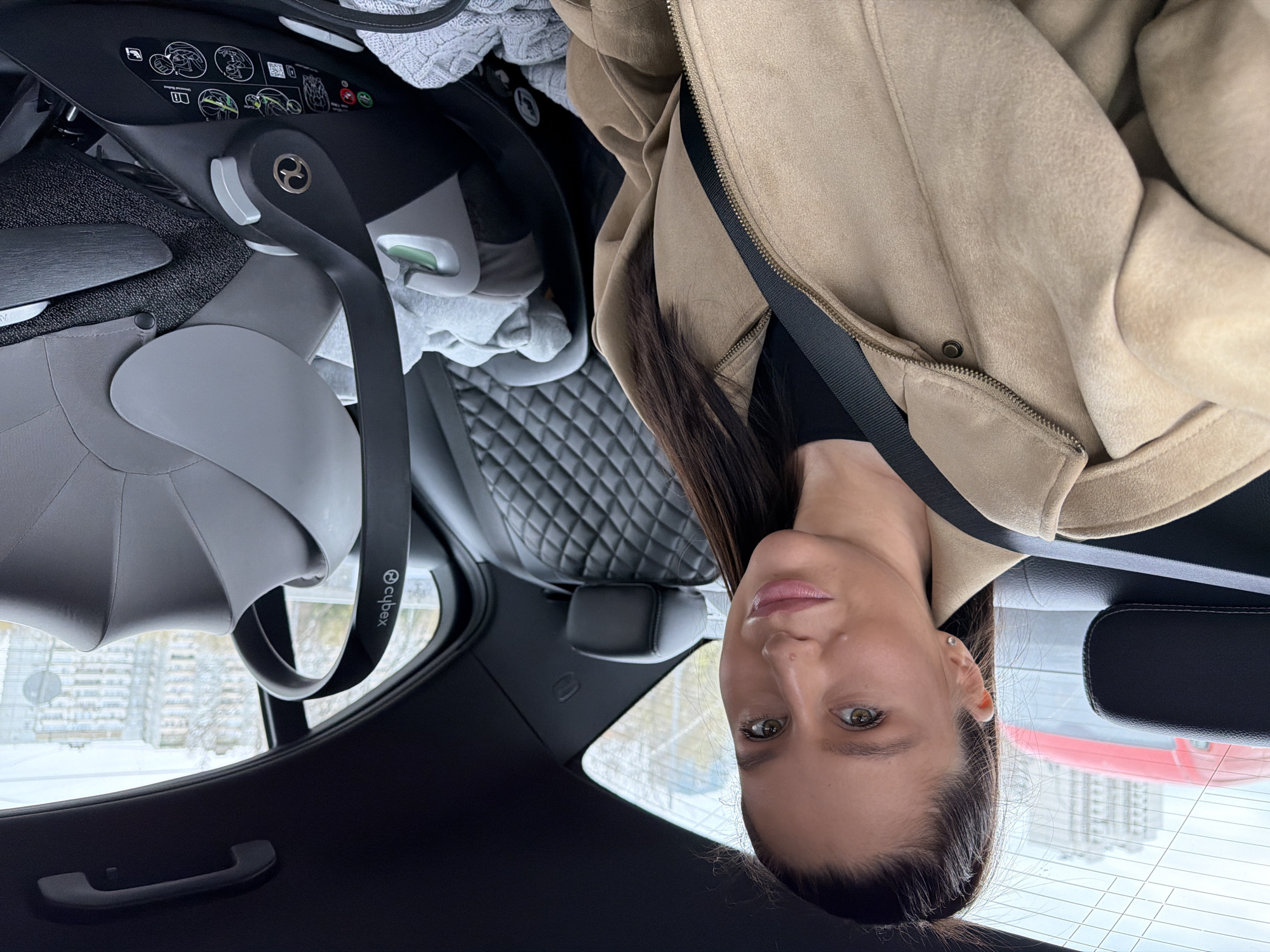 A woman is captured in a close-up, looking directly at the camera from inside a vehicle. She has dark hair, light eyes, and a neutral expression with slightly parted lips. She is wearing a beige or tan jacket, possibly made of suede, over a dark top, with a dark seatbelt strap crossing her chest. A small stud earring is visible in her left ear.
Behind her, in the back seat of the car, is a prominent Cybex baby car seat in shades of gray and black. The brand name "cybex" is clearly visible on its handle. Various instructional and warning symbols and diagrams for installation are printed on the car seat's base. A gray knitted blanket and another lighter gray fabric are draped around the car seat. The car's interior features quilted black leather upholstery on the back seat and dark plastic trim, including a door handle.
The scene appears to be set during daytime, with an overcast sky. Through the car windows, an urban landscape is visible, including buildings and rooftops with hints of snow, suggesting a winter season in Moscow, Russia. The woman appears to be a passenger, possibly taking a selfie, while traveling or waiting in the car.