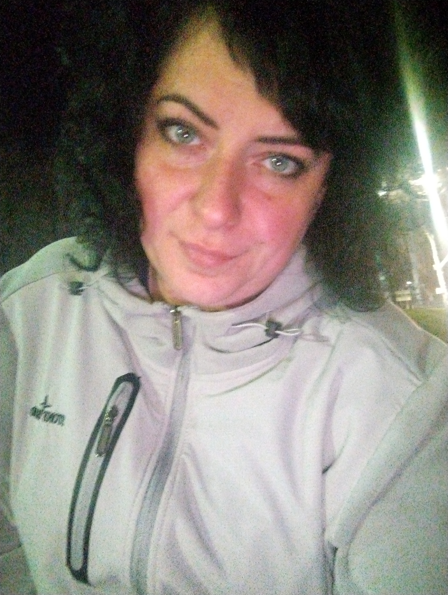 A woman is shown in a selfie. She has dark curly hair and light-colored eyes. She is wearing a light-colored jacket with a zipper. The background is dark, with some light suggesting a city environment. It is claimed to be the city of Votkinsk, Russia, although this cannot be verified. The woman is looking directly at the camera with a slight smile.