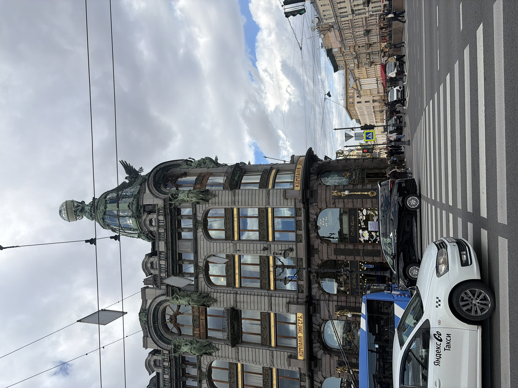 An urban street scene in Saint Petersburg, Russia, is captured under a cloudy daytime sky. The prominent subject is the Singer House (Dom Knigi / House of Books), an ornate Art Nouveau building with a distinctive glass-domed tower, visible on the left side of the frame. The building's facade features large arched windows, decorative sculptures of mythical figures, and gold accents. Visible text on the building includes "КОМПАНИЯ ЗИНГЕРЪ" (Singer Company) and "Cafe René".
In the foreground and extending towards the right, there's a bustling street with a wide zebra crossing. Several cars are visible, including a white Yandex Go taxi with the text "Яндекс Go Такси" printed on its side, and a blue bus. Other vehicles, including a black car, are parked or moving on the street. Numerous pedestrians can be seen on the sidewalks and near the crosswalk, suggesting a busy urban environment. Overhead, various power lines and traffic signals crisscross the sky. In the distance, other historical buildings line the street, characterized by their classical architecture and multiple stories. The overall impression is one of a lively city center on a day with overcast skies.