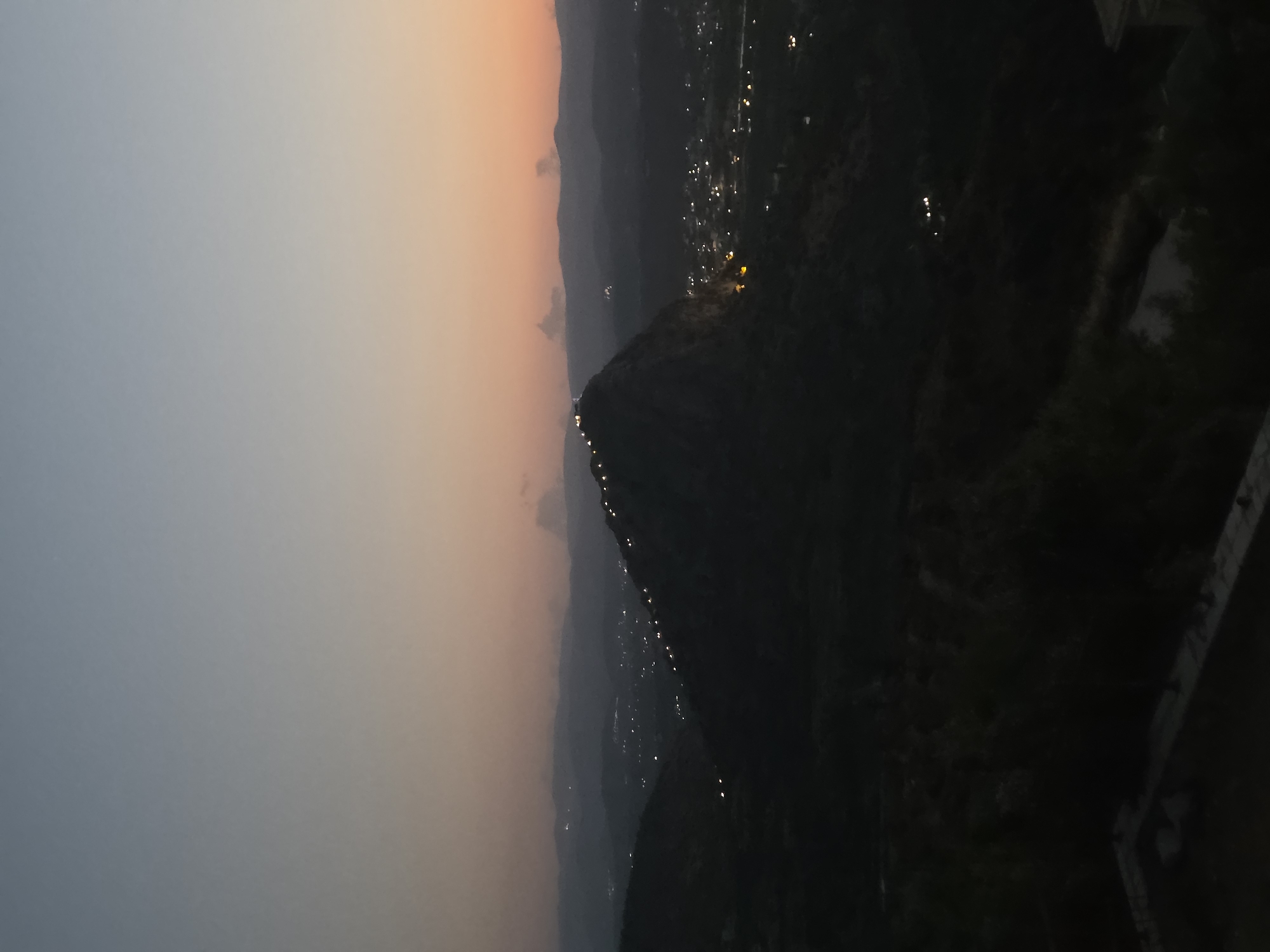The image shows a panoramic view of a mountainous landscape at dusk, with the lights of a town or village visible in the valleys and on the slopes. A prominent, dark, conical mountain peak dominates the center of the frame, with a line of lights illuminating its crest, suggesting a path or structure. The sky exhibits a gradient of colors, transitioning from a deep blue at the top to a soft orange and pink near the horizon, characteristic of sunset. Distant hills and mountains are silhouetted against the fading light, creating layers of depth. The foreground is dark and indistinct, with what appears to be a concrete structure, possibly a balcony or terrace, on the lower right. The scene evokes a sense of tranquility and a remote, elevated perspective overlooking a populated area nestled within the natural terrain. This scene is likely captured from an elevated viewpoint in the Municipal Unit of Asini, Greece, given the characteristic mountainous terrain and the presence of scattered settlements often found in such Greek regions.