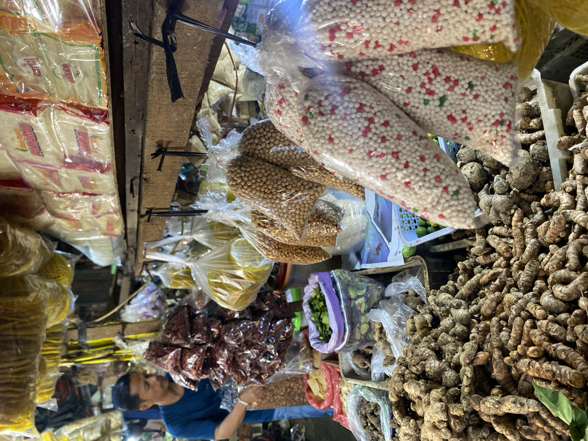 This image captures a scene at a market stall in Cikupa, Indonesia. The stall displays a variety of dried goods and spices. In the foreground, a large pile of ginger roots is visible, with some loose green leaves on the left. To the right of the ginger, there are bags of small white and red beads, possibly decorative candy or spices, and behind them, bags of brown spherical items, likely nuts or seeds. Further back, a person wearing a blue shirt is standing behind a display of dried red chilies in plastic bags. The stall is covered by a wooden structure with hanging items, including yellow circular snacks in plastic bags and other goods wrapped in clear plastic. The environment appears to be an open-air market with natural lighting.