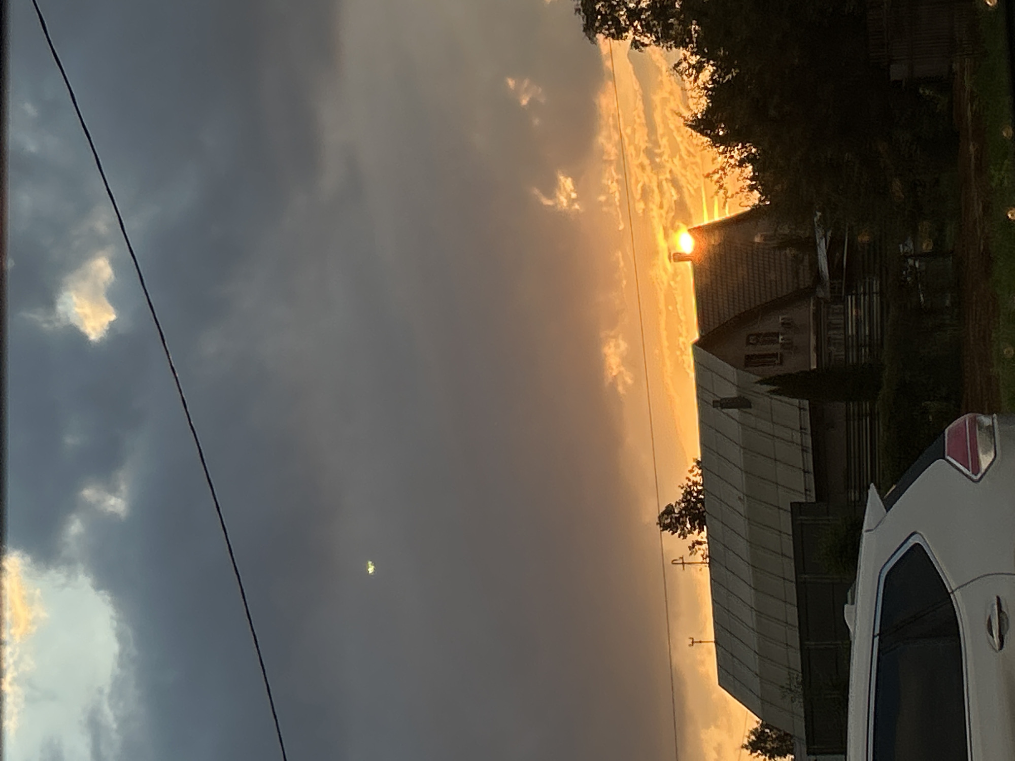 A cloudy sky at sunset is visible over two houses and a white SUV parked in front of them. The sun is setting behind the houses, casting a warm orange glow on the clouds. There is a single power line stretching across the sky. The houses appear to be in a rural or suburban area. There are trees and bushes surrounding the houses. A white SUV is parked in the foreground on the left side of the image. There are no people visible in the image. The scene is likely in Ivanovo, Russia, given the context of the query.