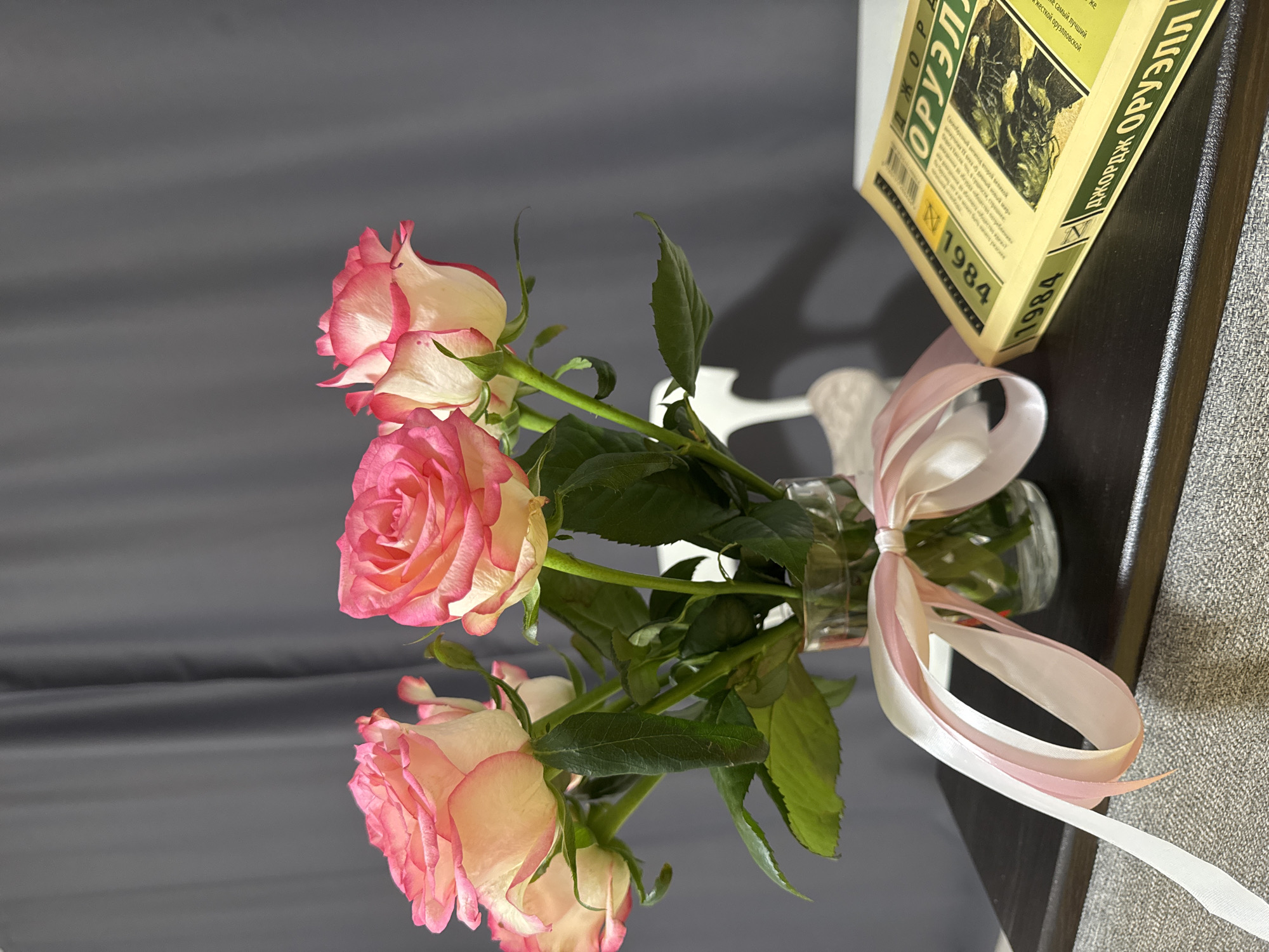 The image shows a bouquet of pink and white roses in a clear glass vase, tied with a pastel ribbon. To the right of the vase is a yellow book with Cyrillic lettering, titled "1984" and authored by "George Orwell" (Джордж Оруэлл). The book is lying on a dark surface, which appears to be a shelf or table, positioned in front of a textured grey curtain. A portion of a white chair is visible behind the roses. The overall scene is indoors, with soft lighting. There is no specific indication of the city of Mariupol, Ukraine.