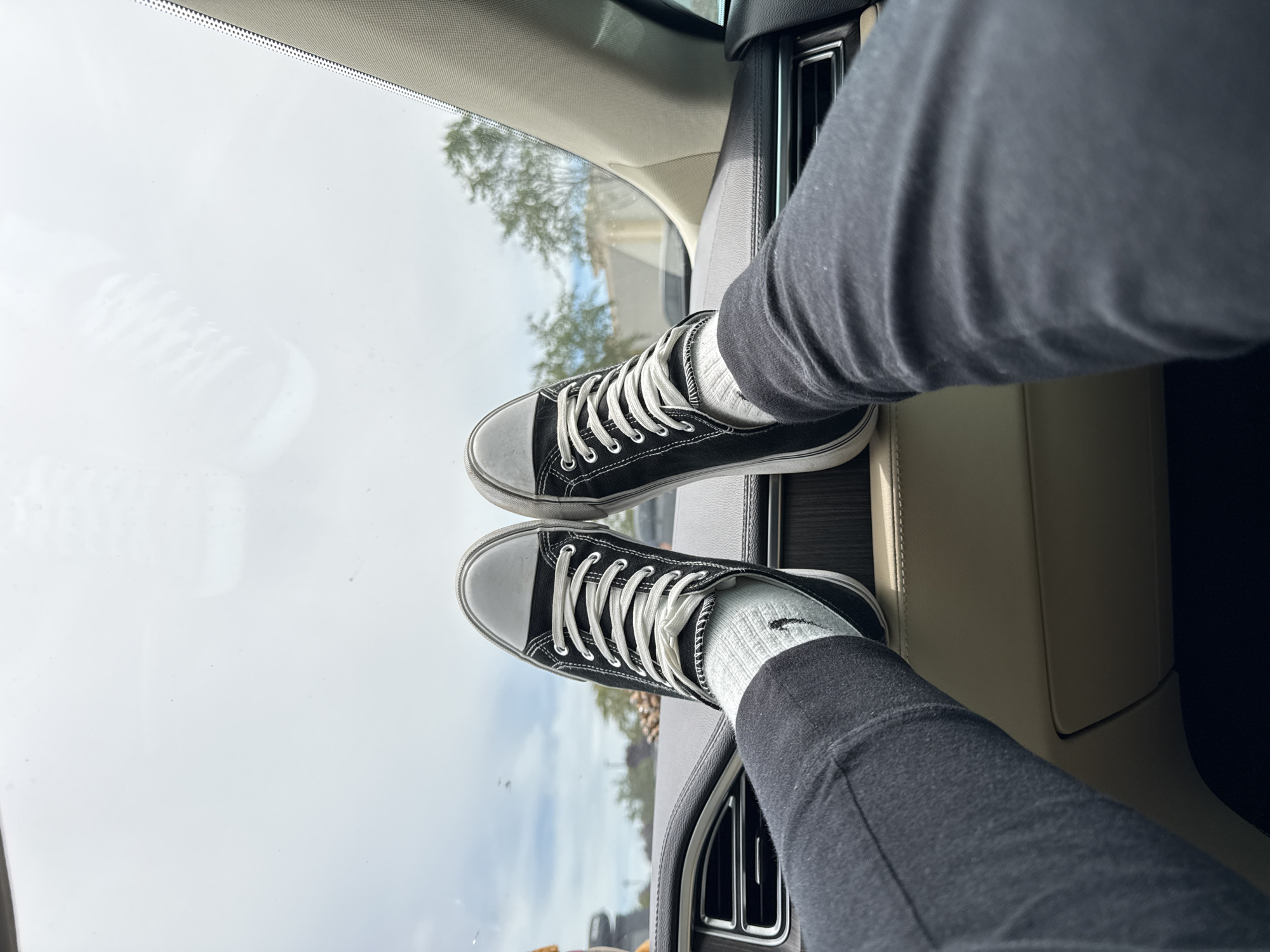 In Lakewood, United States of America, a close-up, first-person perspective shot captures a pair of feet resting on a car's dashboard. The individual's legs, clad in black pants, extend upwards, with feet adorned in black canvas sneakers and white socks. The sneakers have white laces and rubber soles. The car's interior, featuring a dark dashboard with horizontal vents and a beige trim, is visible. Beyond the windshield, a cloudy sky and distant trees can be seen. The scene is static, with no apparent interaction or action.
