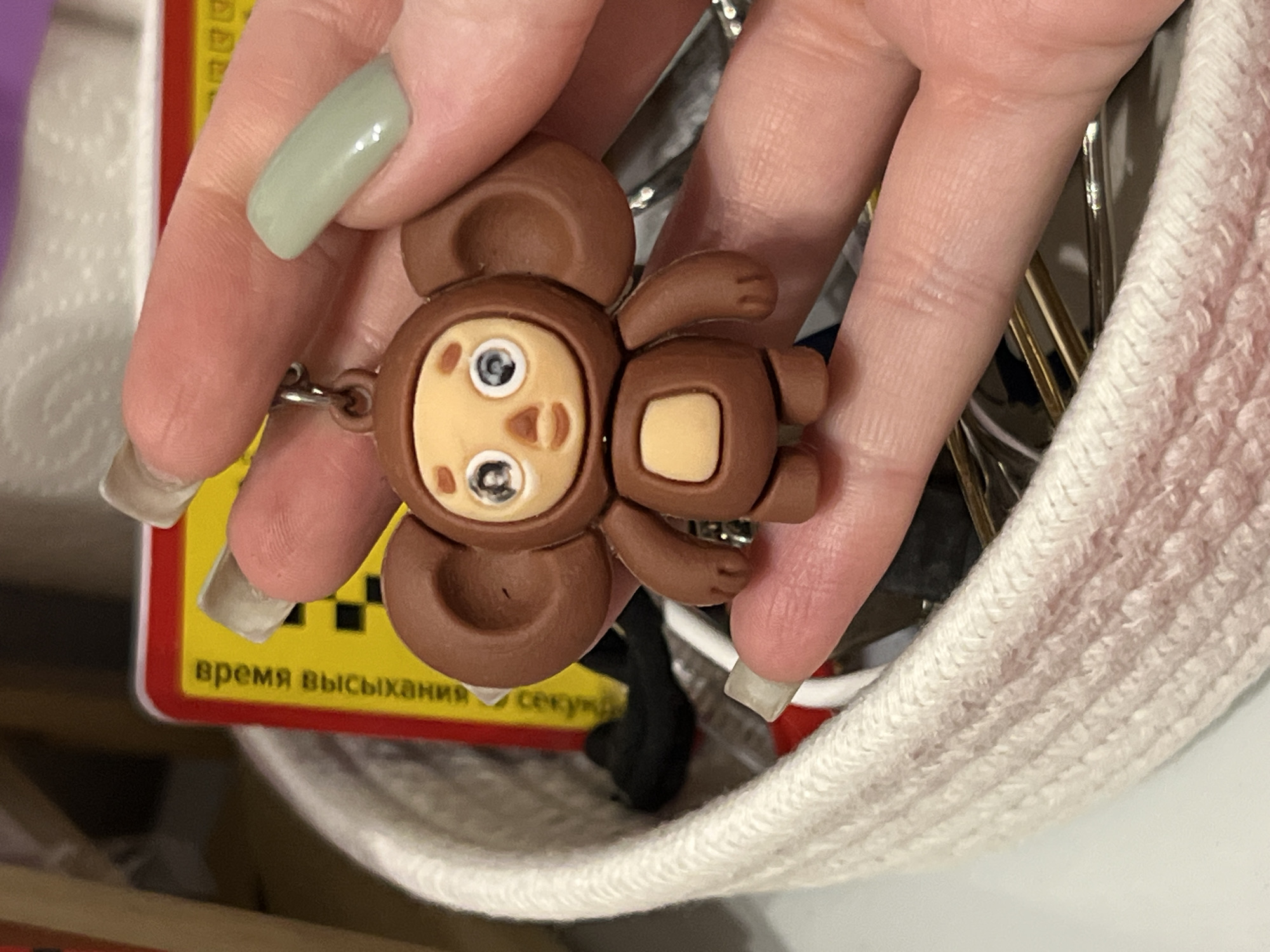 The image shows a keychain in the shape of Cheburashka, a popular Russian cartoon character, being held by a person's hand. The keychain is brown with a light tan belly and face, and large round ears. The Cheburashka's eyes are large and white with black pupils. The hand holding the keychain has painted fingernails, with the index finger sporting a light green polish. The background includes a white woven basket and a yellow card with Russian text that reads "ВРЕМЯ ВЫСЫХАНИЯ" (drying time). The setting appears to be indoors, possibly a desk or craft area, and there is no indication of the specific location within Kazan, Russia.