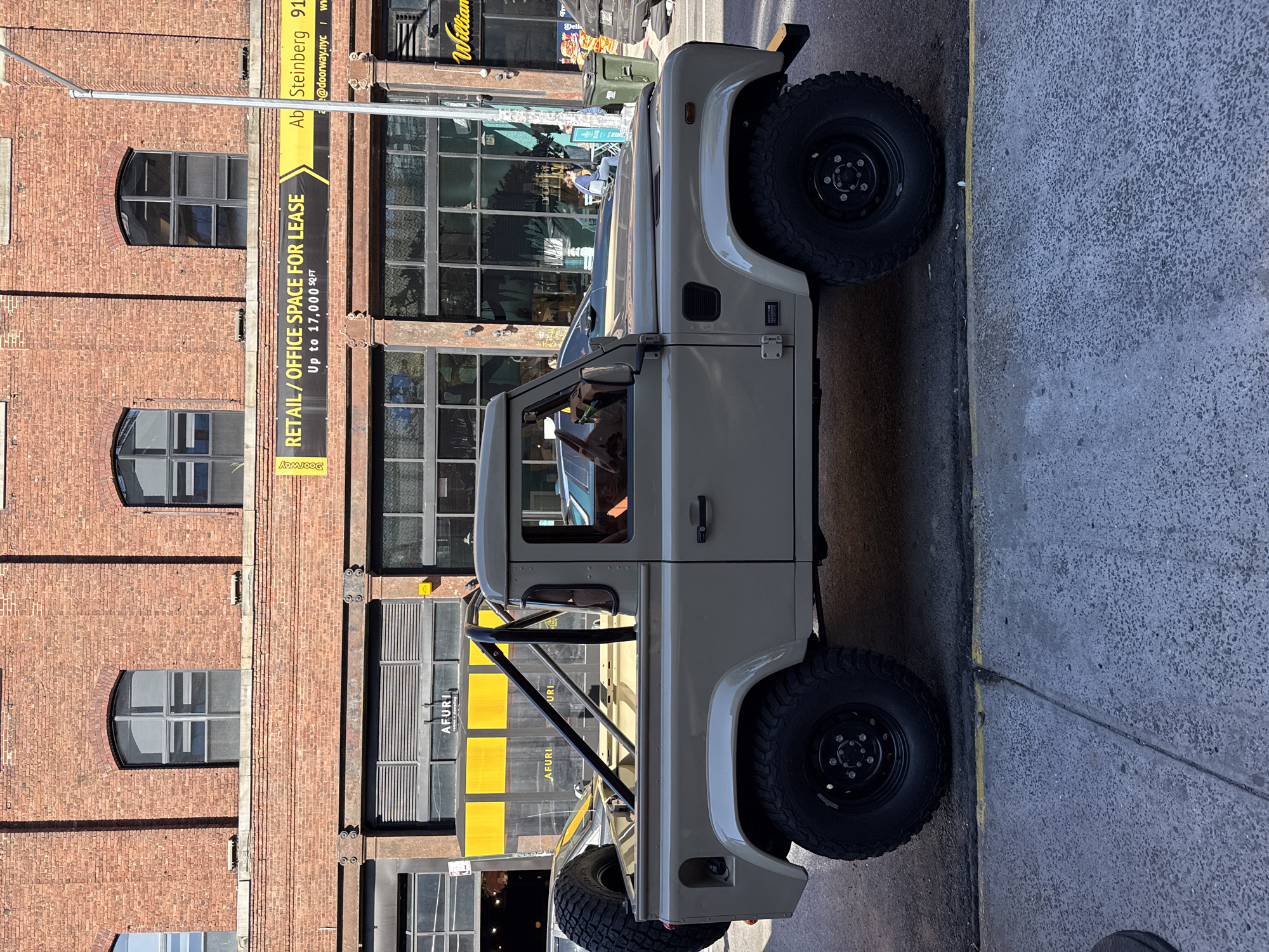 The image captures a beige off-road vehicle, likely a Land Rover Defender, parked on a street in New York City, United States of America. The vehicle's exterior is detailed with visible black tires and a black roll cage extending above the vehicle's rear.
Behind the vehicle, the urban environment is evident in the brick architecture of the buildings. A banner advertising "RETAIL/OFFICE SPACE FOR LEASE" is displayed on the building. A restaurant called "AFURI" is also visible on the storefront. The setting suggests a commercial area.