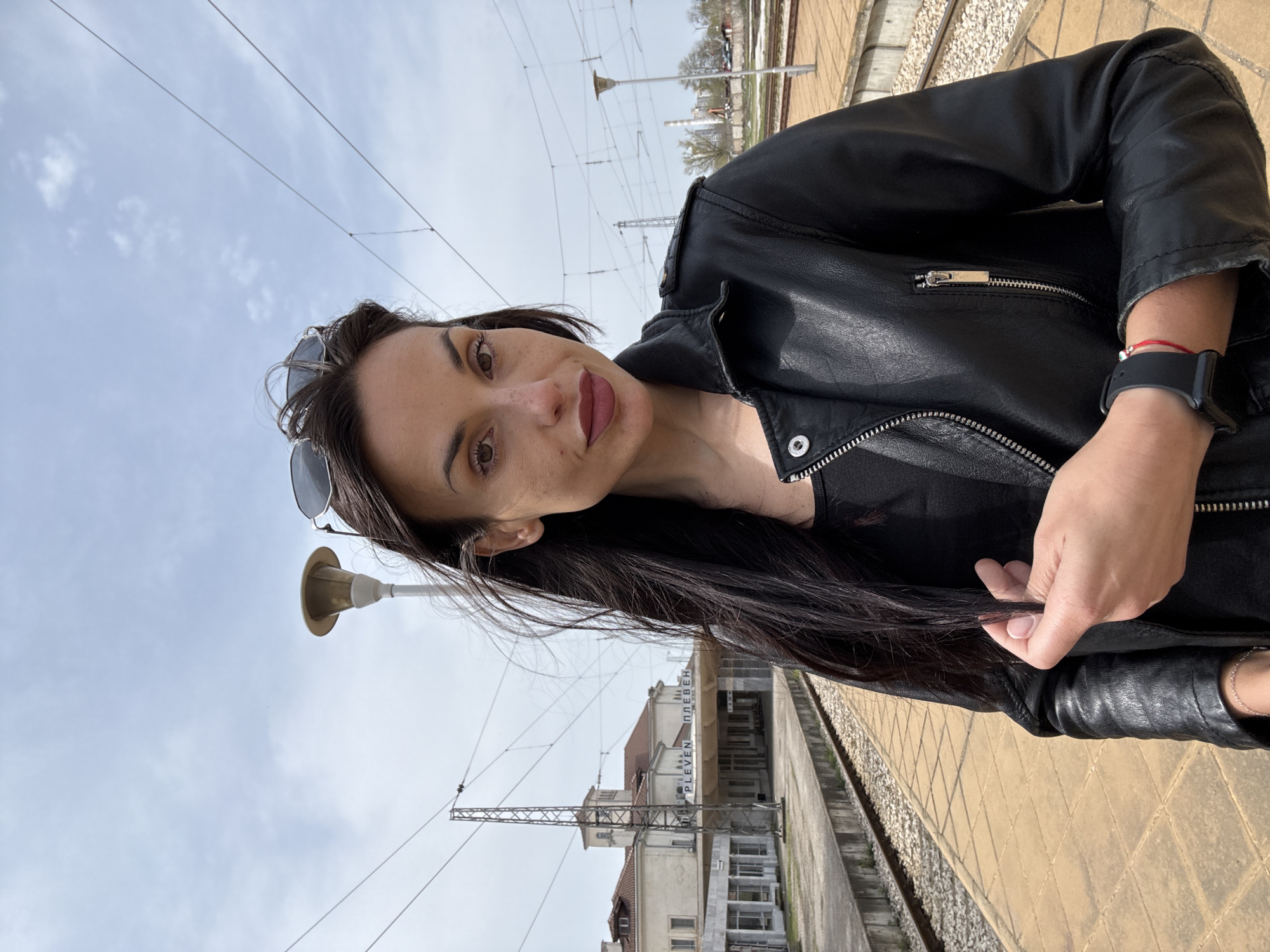 A woman is standing on a train platform in Pleven, Bulgaria, facing the camera and tilted to the left. She is wearing a black leather jacket and sunglasses are perched on her head. She is holding a strand of her long, dark hair. Behind her, a building with the word "ПЛЕВЕН" (Pleven) on it can be seen, indicating the location is Pleven train station. The platform is paved with beige tiles. Train tracks run alongside the platform. Above, there are power lines and support structures for the railway. The sky is overcast.