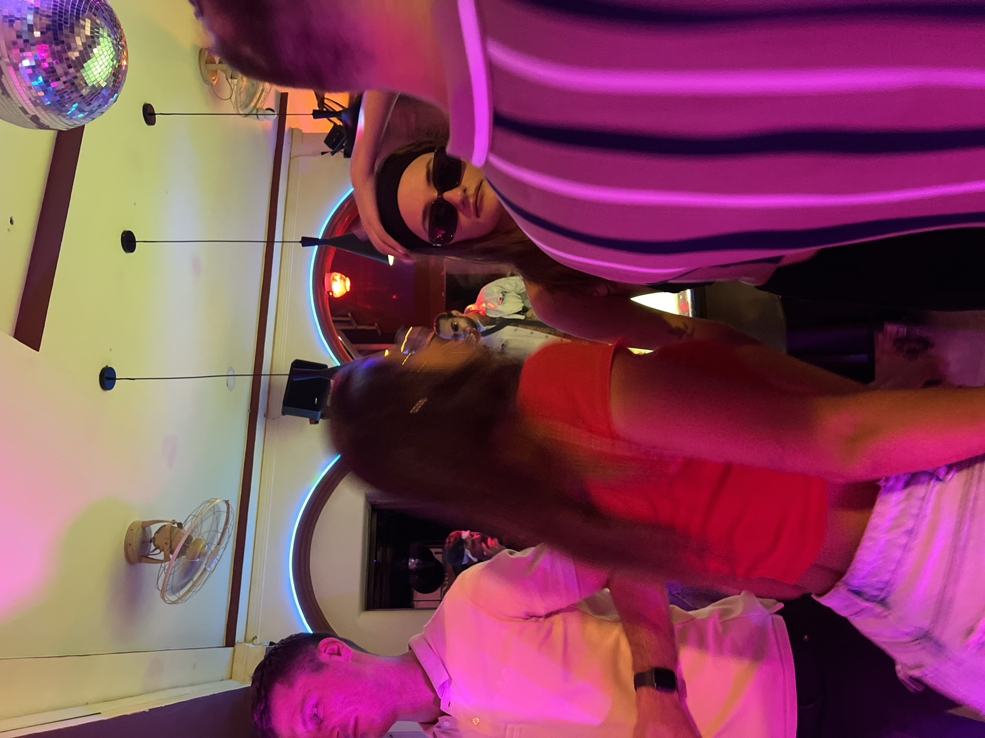 In Nha Trang Ward, Vietnam, an indoor scene is captured at an indiscernible location. Three individuals are visible, partially obscured within the frame. One person is wearing a horizontally striped top, another is clad in a red shirt, and the third person is wearing a white shirt. A woman is wearing sunglasses and a headband. The environment includes a disco ball, wall-mounted fans, and neon-lit circular fixtures.