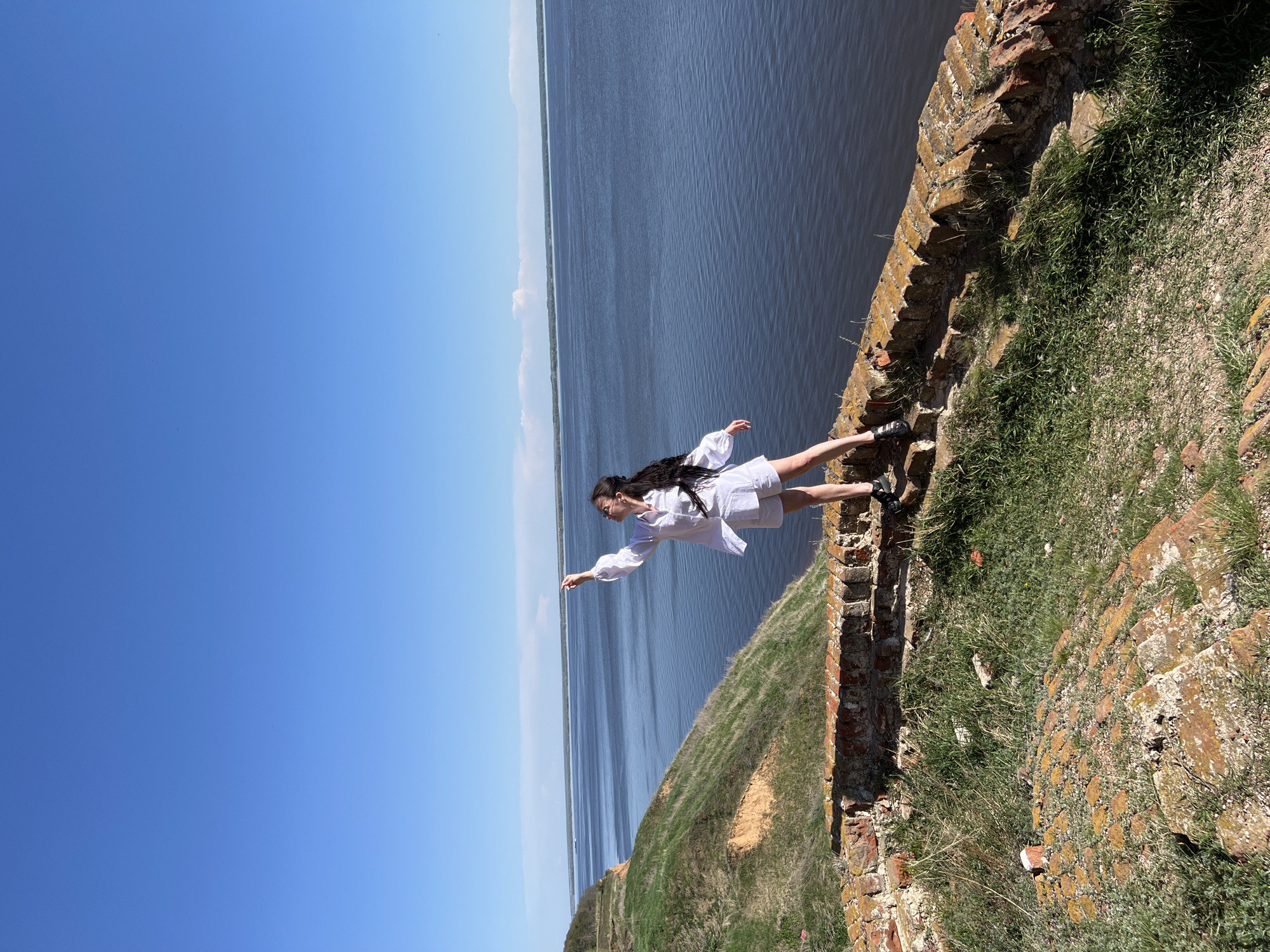 A medium shot angled with a woman in a white outfit standing on a wall by a body of water under a blue sky.
The woman stands on a crumbling brick wall with vegetation growing on it. She wears a long-sleeved white top and white shorts, with dark footwear. Her dark hair flows down her back. She has one arm outstretched, pointing towards the water. The water stretches to the horizon, where a thin line of green land is visible. The sky is a clear, light blue.
Based on geolocation, the location is near Шуран, Russia.