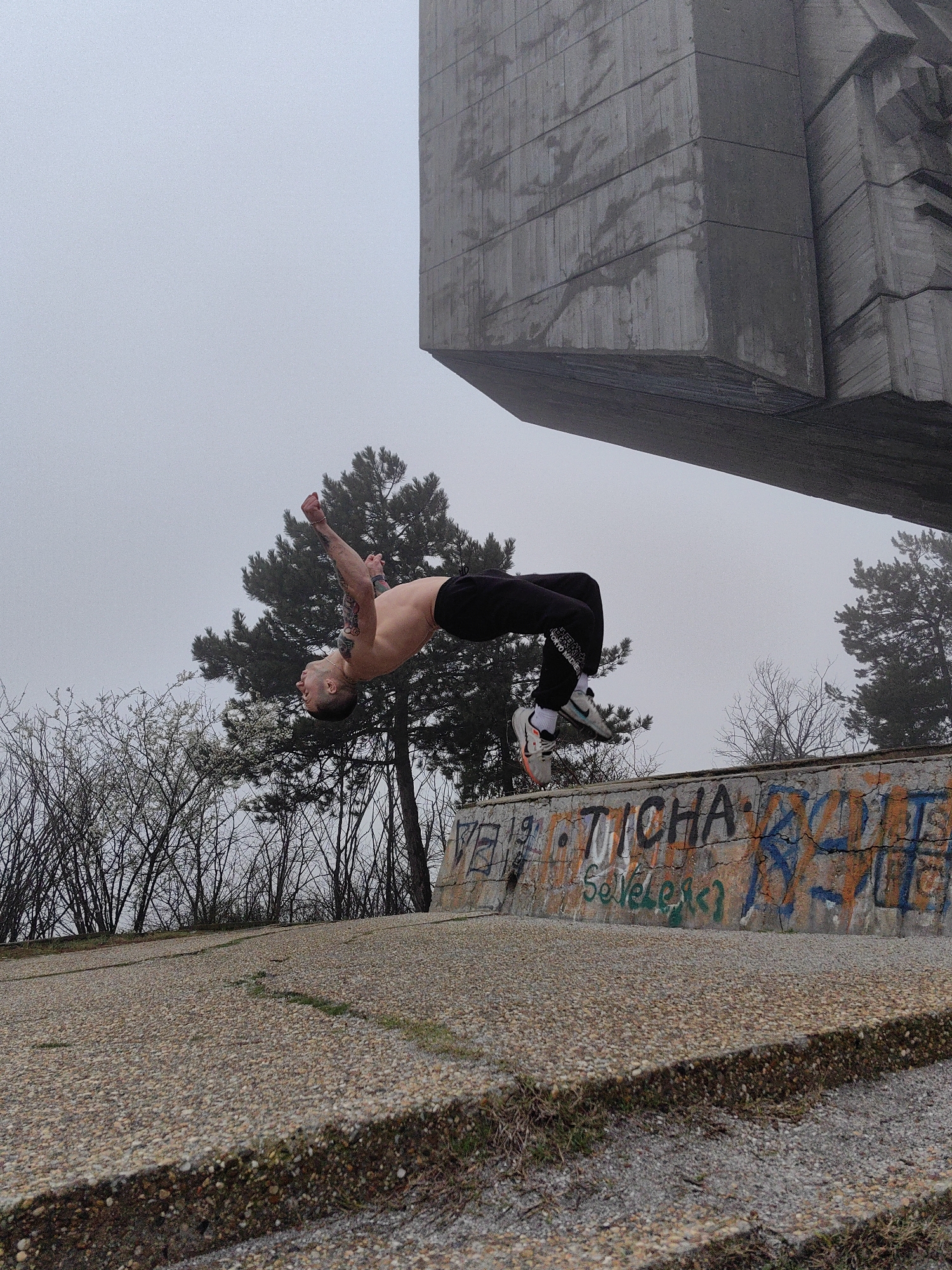 A shirtless man wearing black pants and sneakers performs a backflip in front of a concrete monument with graffiti. The location is likely in Varna, Bulgaria, given the style of monument and graffiti. The sky is overcast. Trees surround the monument. A concrete walkway or plaza with visible seams and edges leads to the monument.