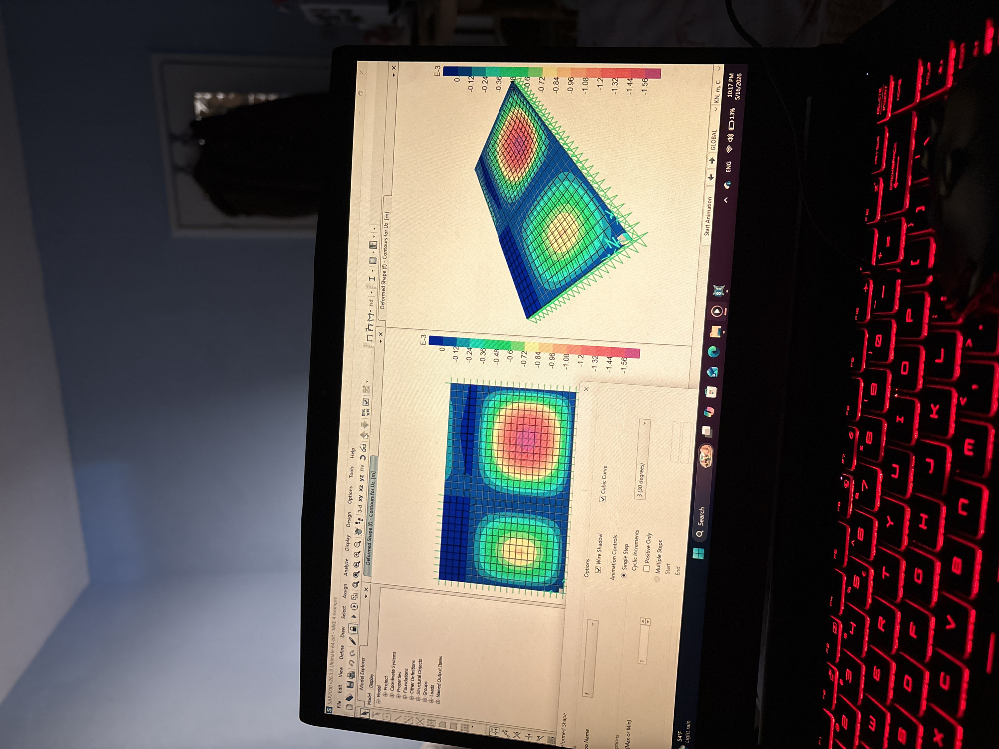 A laptop screen displays an image of a structural analysis software program, specifically SAP2000 v26.3.0. The software interface is visible, including menu options like "File," "Edit," "View," and "Define," as well as icons related to model display and analysis. The main window shows a deformed shape analysis, with a color-coded contour plot representing the displacement Uz (likely the Z-axis displacement) of a structural element. The color scale ranges from 0 to -1.56 E-3 (meters). An additional "Options" menu displays settings such as "Wire Shadow" and "Cubic Curve" with a 3 (30 degrees) specification.
The laptop itself has a black chassis and a red-illuminated keyboard. The keyboard layout is visible. On the lower right of the screen, system information indicates 10:17 PM on May 16, 2026. The power is at 13%.
The surroundings of the laptop are dark. A partially visible framed image is on the wall behind the laptop.
The location of the scene is not evident from the image. Sofia, Bulgaria cannot be determined.