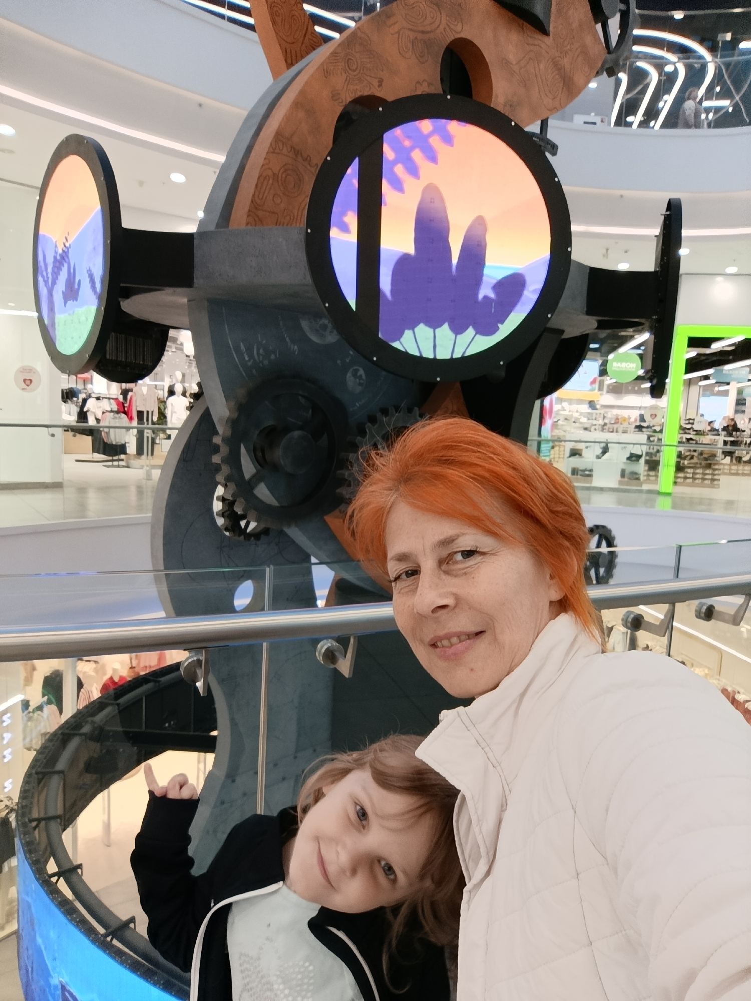 Inside the Aviapark shopping mall in Moscow, Russia, an adult woman with red hair and a white jacket is posing for a selfie with a young girl wearing a black jacket, in front of a large, mechanical art installation resembling a giant telescope. The installation has circular screens displaying animated scenes. The scene includes retail stores in the background.