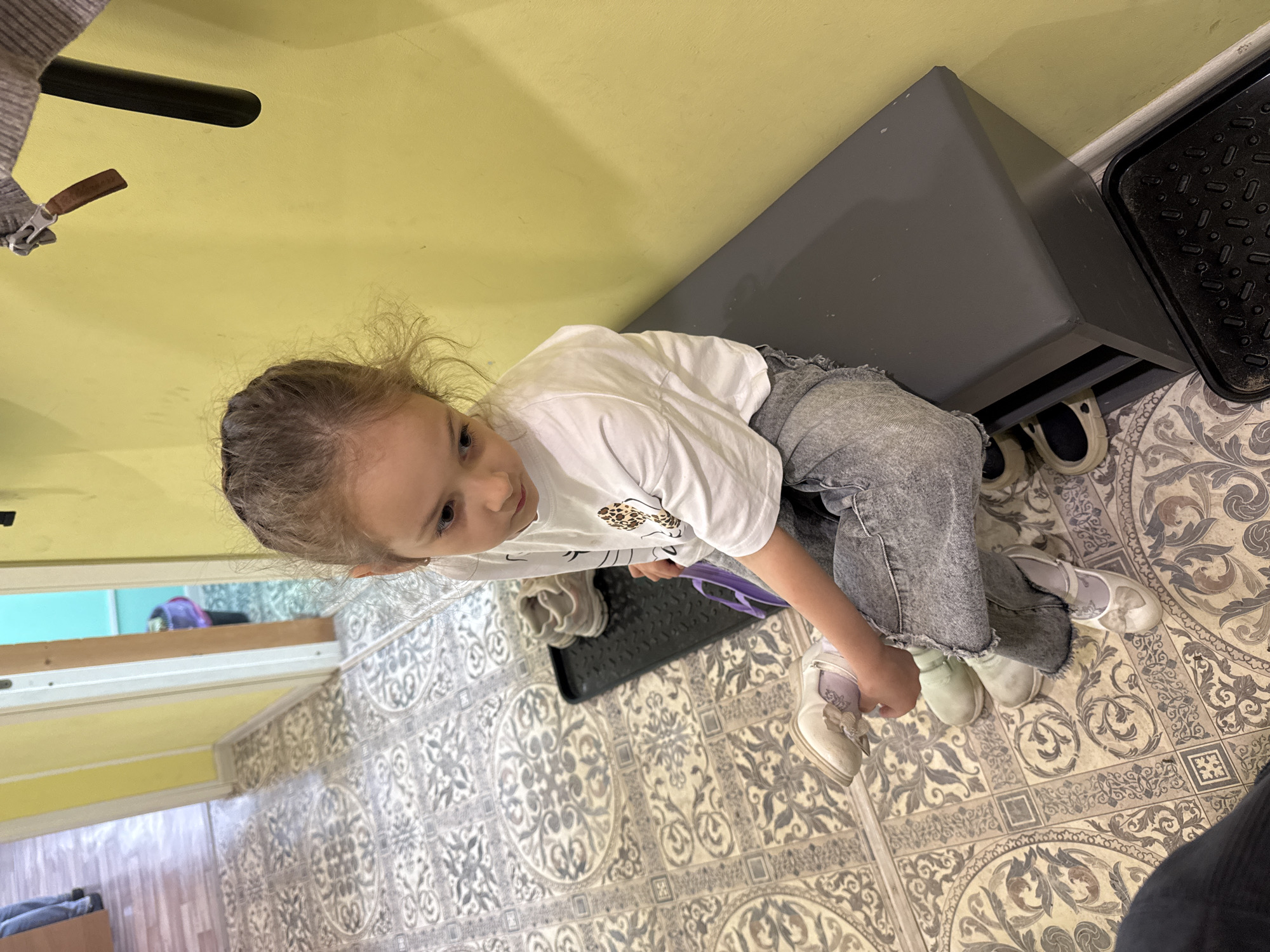 A young, light-skinned girl with blonde hair is sitting on a gray storage bench in an indoor space. She wears a white t-shirt with a printed design, gray jeans, and a pair of mismatched shoes, one off-white and one light green. The flooring is tiled in a complex, patterned gray and beige design. A black metal tray sits on the floor near the bench. The wall is light yellow. Based on the available information, I cannot confirm the exact location as Shevlyakovo, Russia. There is nothing in the image that I can use to confirm that location.