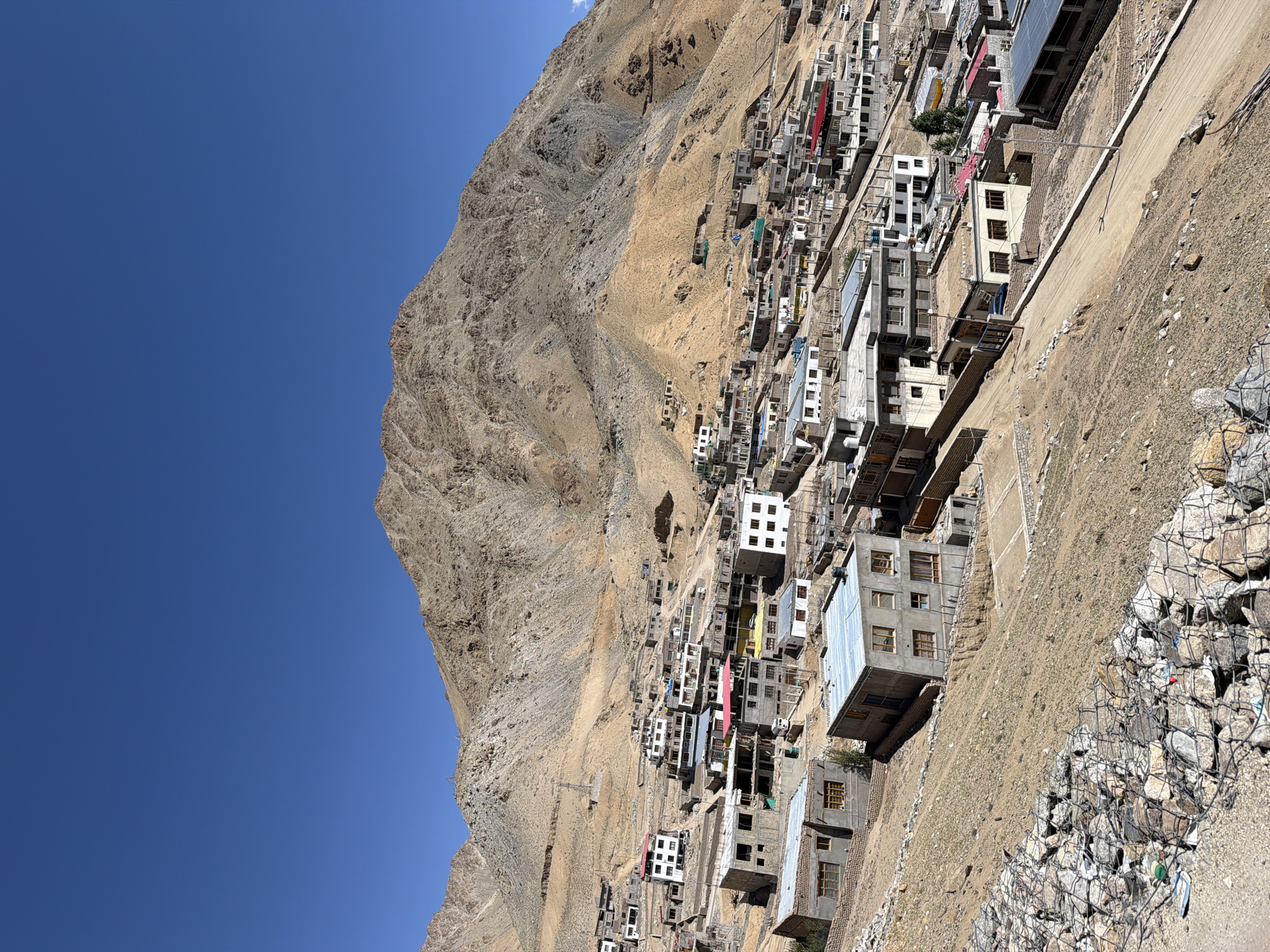 The image depicts a town nestled in a mountainous landscape, characteristic of Leh, India. The foreground features a steep, dry slope covered in gravel and rocks contained by wire mesh. Above this, a cluster of buildings, varying in size and architectural style, are densely packed together, ascending the hillside. Many structures appear to be constructed from concrete and brick, with some featuring flat roofs and multiple stories. Signs of ongoing construction are visible on several buildings. A dirt road winds through the settlement, with a few vehicles parked or driving. The backdrop is dominated by a large, arid mountain range with rugged, textured slopes under a clear, vibrant blue sky. The overall scene conveys a sense of a remote, high-altitude settlement in a dry, mountainous region.