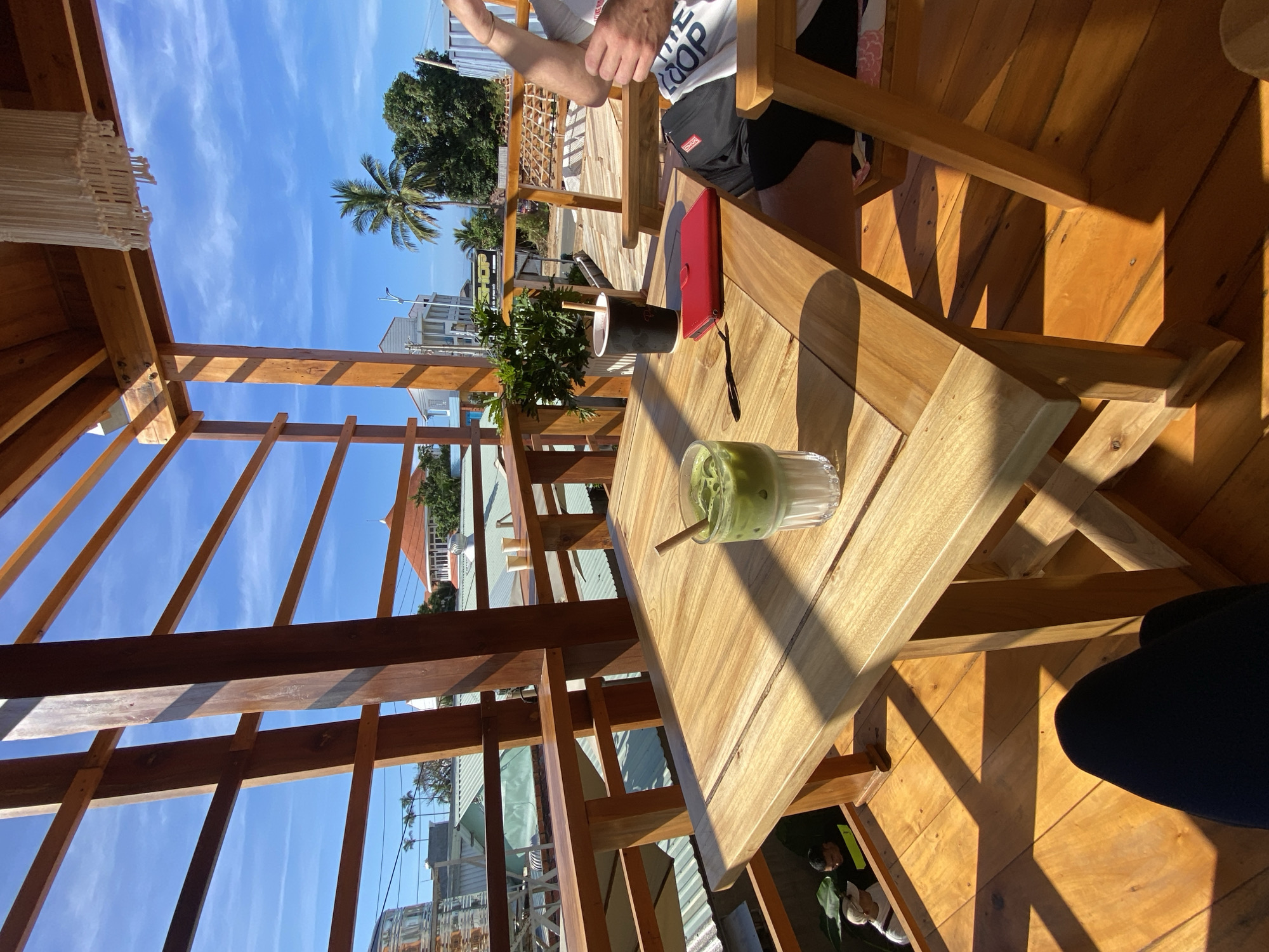 An outdoor cafe in Mũi Né, Vietnam, with a view of buildings and palm trees under a clear blue sky. A wooden table in the foreground holds a glass of green matcha latte with ice and a cinnamon stick, a black cup with a straw, and a red wallet. A person's legs in black pants are visible in the lower foreground. In the background, two men are standing near some plants. The cafe has wooden structures and railings, with sunlight casting geometric shadows on the wooden floor and table.