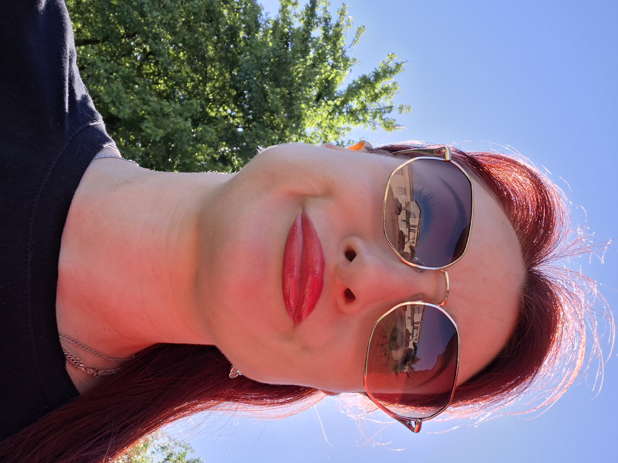 A low-angle portrait shows a woman with fair skin, wearing gold-rimmed sunglasses that reflect buildings. She has red lipstick and red hair, and she is wearing a black top with a silver necklace. A tree and a blue sky are visible in the background.