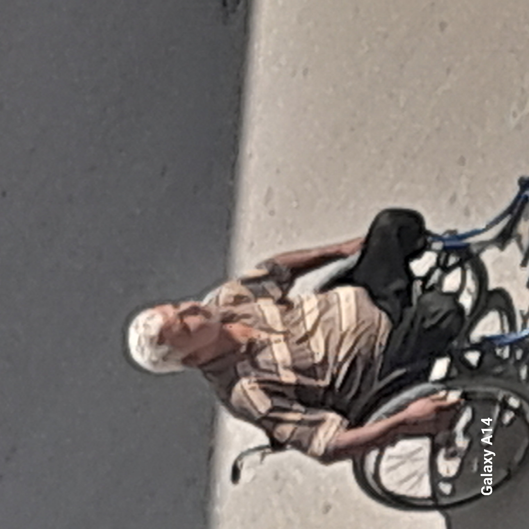 An elderly man with white hair, wearing a striped shirt and dark pants, sits in a wheelchair on a concrete surface. The wheelchair is black with large spoked wheels. The man is looking to his left. The background is a plain, gray, textured surface that appears to be a wall. The lighting suggests it is daytime, with shadows indicating sunlight. A watermark "Galaxy A14" is visible in the lower left corner of the image. The scene is consistent with an outdoor urban environment in Khan Yunis, Palestinian Territories.