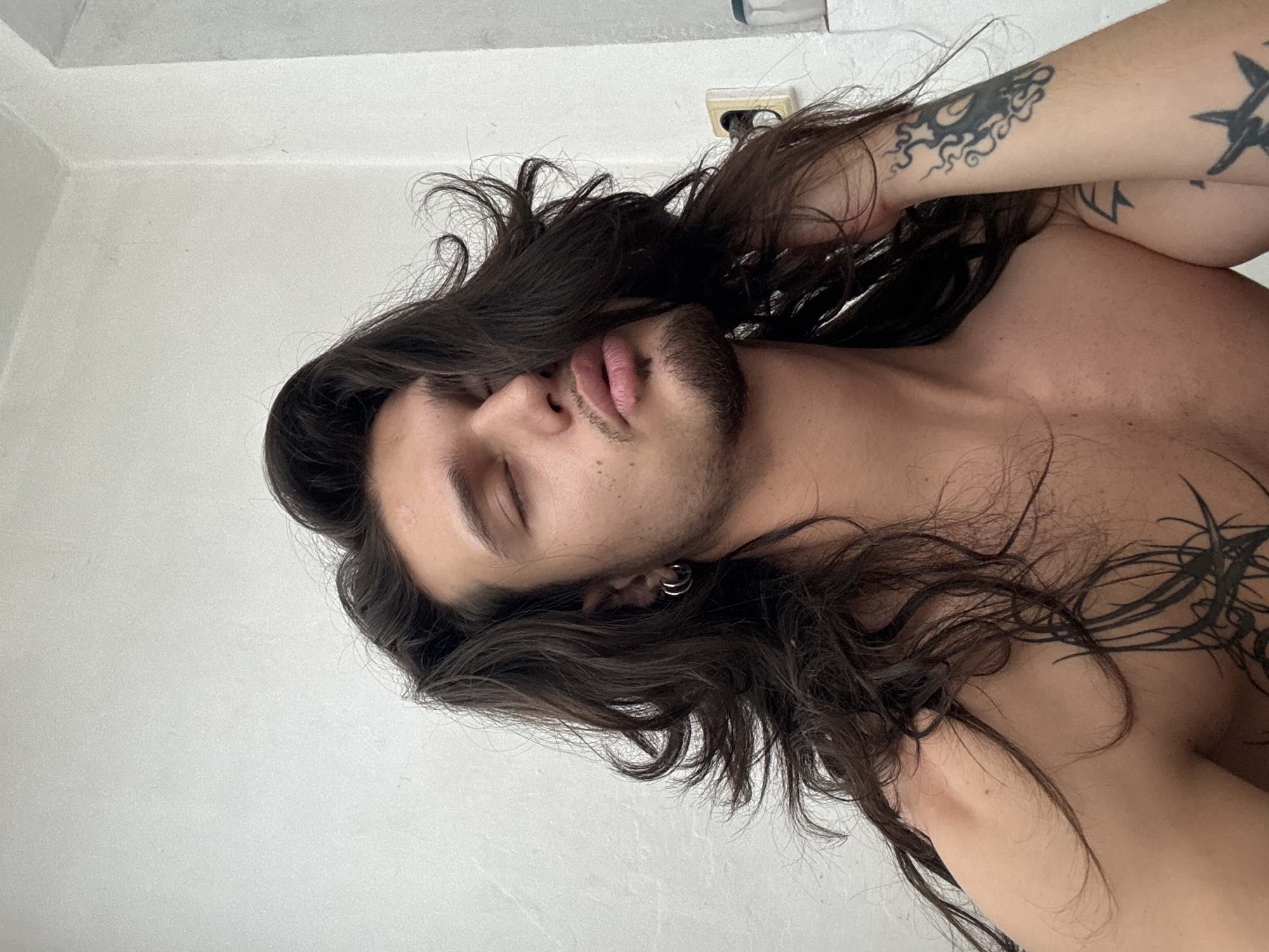A young person with long, dark, wavy hair is shown from the chest up, with their eyes closed and head tilted back. They are wearing a silver hoop earring in their left ear and have a small goatee. Tattoos are visible on their chest and right arm. The background is a plain white wall, with an electrical outlet visible on the right. The lighting appears to be natural, coming from the front and slightly to the left. The image is a close-up, focusing on the person's face and upper body. There is no information available to determine the location in the city of Sofia, Bulgaria.