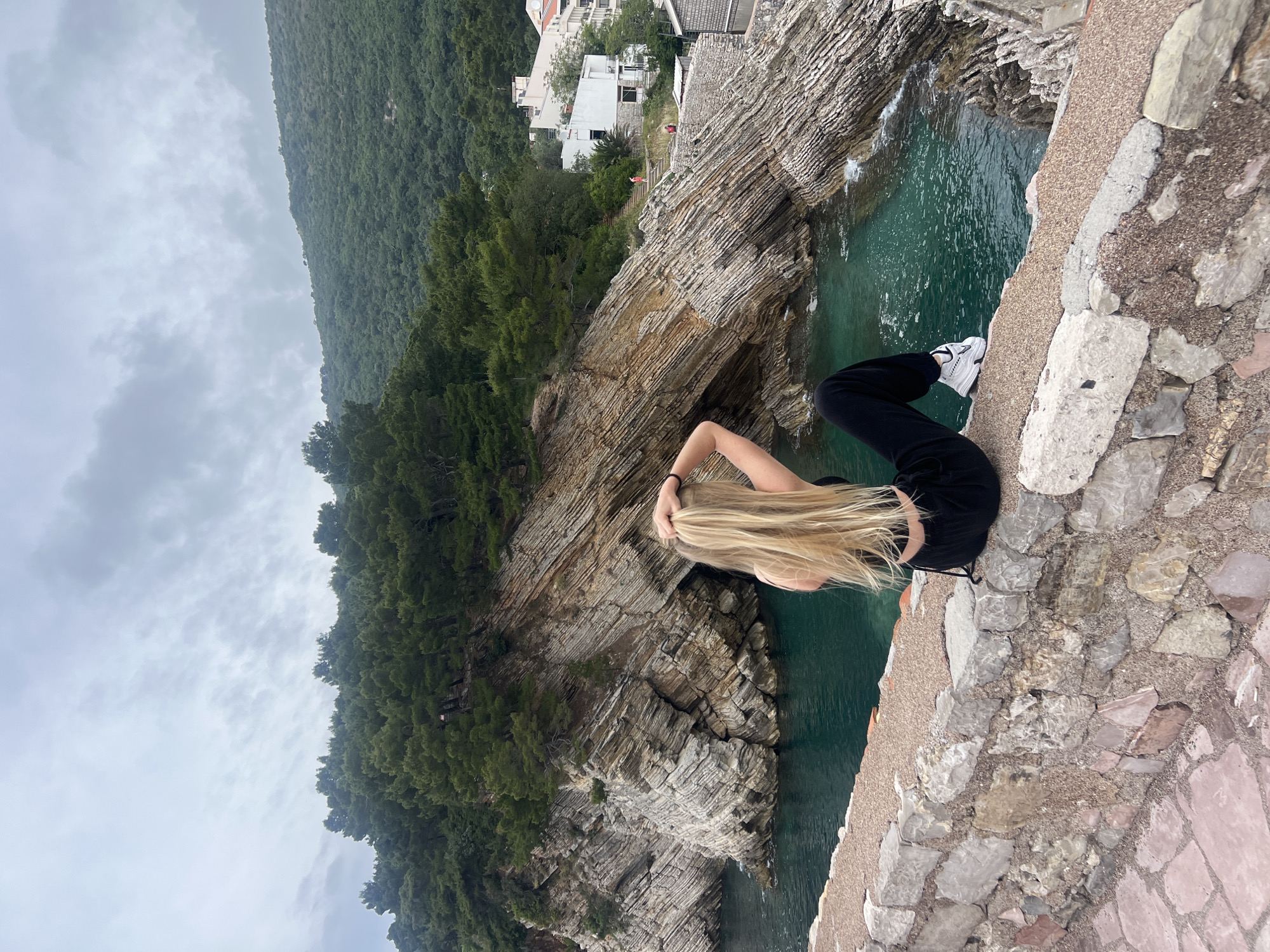 A vertical eye-level shot depicts a blonde-haired woman seated on a stone wall. The wall consists of various-sized stones of different colors, predominantly grey and tan. She is wearing black pants and white sneakers, facing away from the camera. Her right hand is raised, seemingly touching her hair.
Behind her, the landscape includes layered rock formations that plunge into the sea, which is a vibrant turquoise color. A hillside covered in green trees is visible. Further back, there are buildings, suggesting the outskirts of a town. The sky is overcast with grey clouds. The location is Petrovac centar, Montenegro.