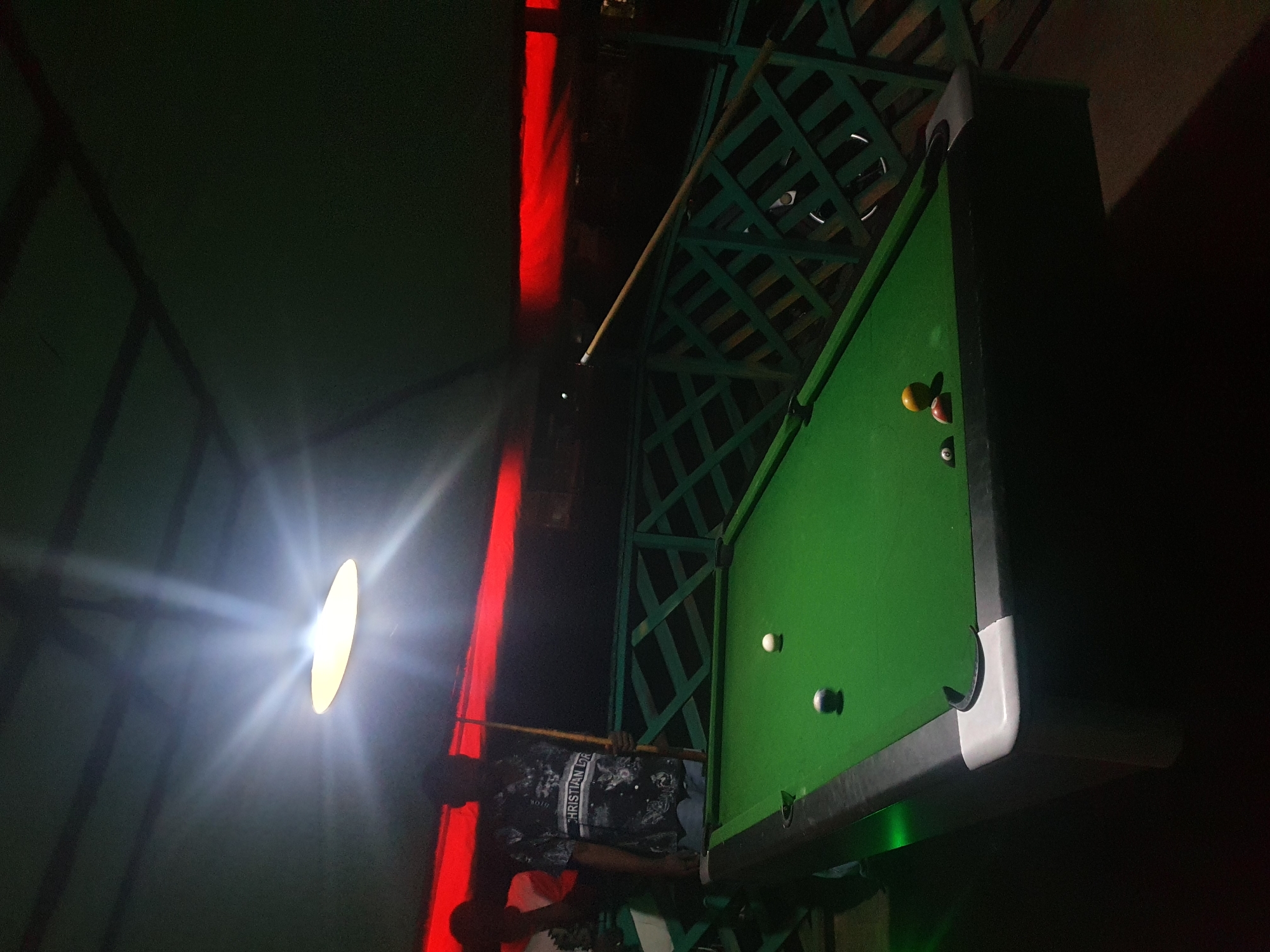 The media file depicts a dimly lit indoor scene, likely a bar or recreation area, with a focus on a billiard table. A person, wearing a black and white patterned shirt with visible text "CHRISTIAN Dior" and "MOTO," is positioned behind the billiard table, holding a cue stick. They appear to be playing or about to play a game of billiards. Several billiard balls are visible on the green felt surface of the table.
The scene is illuminated by a bright, diffused light source on the left, casting strong rays and creating a lens flare effect. A vertical band of red light is prominent on the left side of the image, suggesting ambient lighting or decorative elements. The background is dark, with hints of green lattice-like structures above the billiard table, possibly part of the ceiling or a decorative overhead feature. The overall mood is atmospheric and somewhat moody, given the low light and strong contrasts. There are no clear indicators of the time of day or weather conditions, but the indoor setting suggests it could be anytime. The location is Lokoja, Nigeria, which might imply a specific cultural context for the establishment.