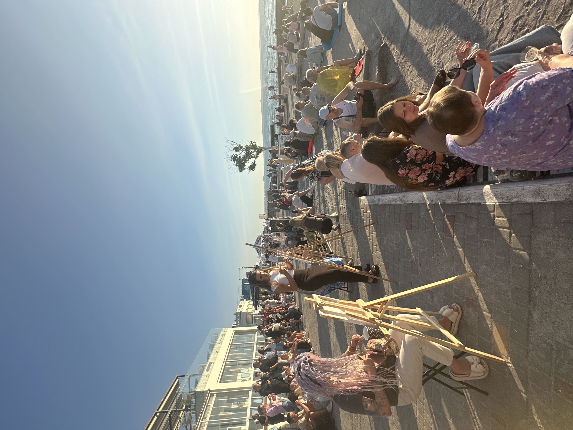 The image depicts an outdoor gathering of people, likely a public event, on a paved promenade adjacent to a body of water, consistent with a seaside location in Odesa, Ukraine. The scene is bathed in the warm, late afternoon sunlight, casting long shadows.
In the foreground, on the left, a woman with brightly colored, braided hair is seated on a stool in front of an easel, appearing to be painting or drawing. She is wearing white pants and light-colored slippers with large bows. Nearby, another woman, dressed in a white top and black skirt, stands beside an easel, looking down at something on it, potentially also engaged in art.
Further into the scene, a crowd of people are seated on the ground or on low steps, facing the water. They are casually dressed, suggesting a relaxed atmosphere. Some are in small groups, conversing, while others appear to be observing the activities or simply enjoying the environment.
Behind the seated crowd, a building with large windows and an outdoor seating area is visible. Further in the distance, the sea stretches out, with a few boats or ships on the horizon. A lone, slender tree stands on the promenade, adding a natural element to the urban setting.
The overall impression is one of a community event, possibly an art exhibition or a public gathering, taking place along the waterfront of Odesa. The clear blue sky and the golden light suggest a pleasant day.
