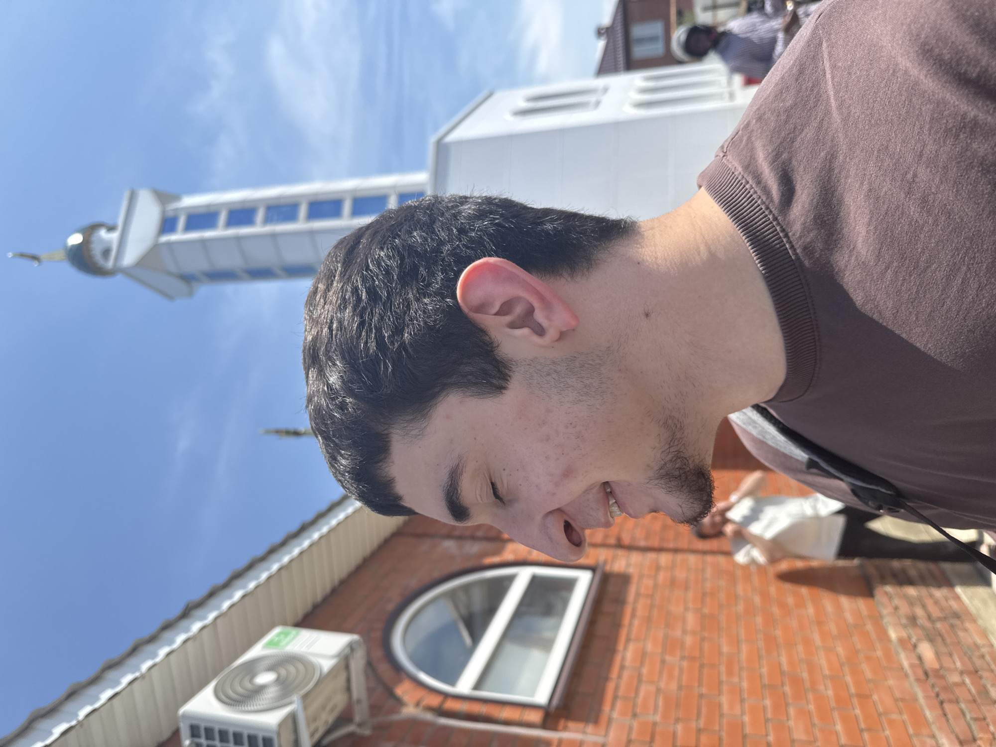 A young man with dark hair and a goatee is shown in profile, smiling with his eyes closed. He is wearing a brown t-shirt and a dark strap across his shoulder. Behind him, a brick building with a white-trimmed arched window and an air conditioning unit is visible. Further in the background, a tall, white minaret with blue windows and a green dome, characteristic of a mosque, stands against a clear blue sky. Two other individuals are partially visible in the background: one in a white shirt and dark pants, and another wearing a helmet. The scene appears to be outdoors in the city of Novaya Adygeya, Russia, under bright daylight.