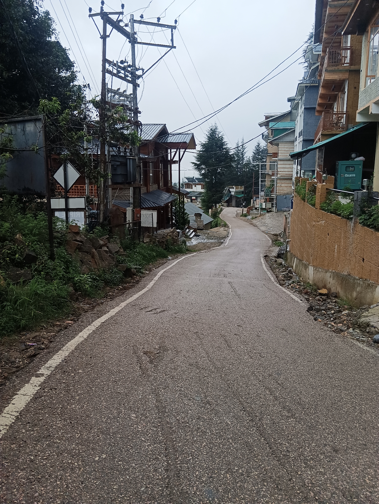 The image depicts a paved road in a hilly, possibly rural or semi-urban area. The road is wet, suggesting recent rain or damp conditions, and has a white line painted along its left edge. On the left side of the road, there is a slope covered with lush green vegetation, rocks, and trees. A large electrical pole with numerous wires and a transformer is prominently visible. Behind the vegetation, a structure with a dark metal roof and wooden elements is present. Further up the slope, another building with a similar dark roof can be seen.
On the right side of the road, a series of multi-story buildings are lined up along the street. These buildings appear to be hotels or residences, characterized by wooden balconies, windows, and pitched roofs. One building has a textured brown wall facing the road. A green generator unit is visible near the buildings. The overall environment suggests a location in a mountainous region, possibly the Himalayas, with construction typical of such areas. The sky is overcast, contributing to a subdued lighting condition. The image is a vertical shot looking up a winding road.