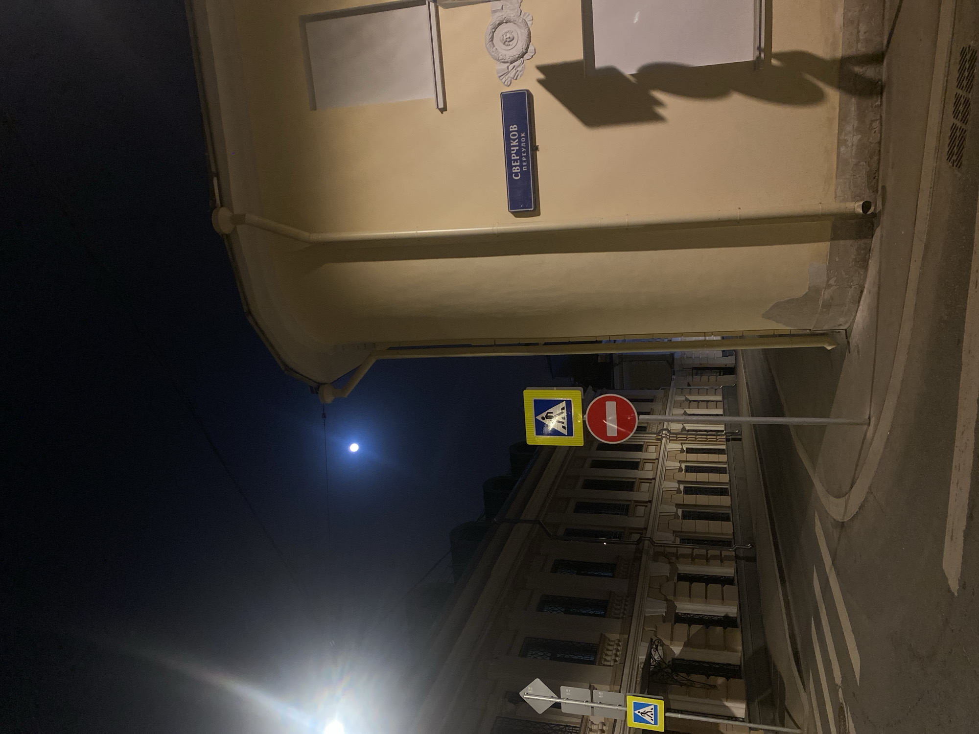 The image depicts a night scene in Moscow, Russia. A sign on a building reads "Сверчков переулок" (Sverchkov pereulok, which translates to Sverchkov Lane). The environment is urban, with buildings lining the street. A crosswalk is visible, indicated by pedestrian crossing signs and road markings. A bright light source illuminates the lower part of the scene, and the moon is visible in the dark sky above. There are traffic signs indicating a pedestrian crossing and a stop sign.