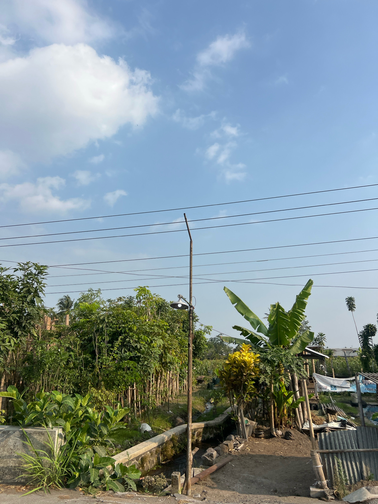 The photograph captures an outdoor scene on a sunny day in Pakem, Indonesia. The sky is a clear blue with scattered white clouds. In the foreground, there is a dirt path leading towards a small structure with a corrugated metal roof, partially obscured by vegetation. Several power lines run horizontally across the upper portion of the image. To the left of the path, there is a concrete structure and lush green plants. A street lamp with a light fixture is visible on the right side of the path. The background consists of dense green foliage, including banana trees, and a small canal. In the distance, there are indications of other buildings and a banner with text. The overall impression is of a rural or semi-rural environment.