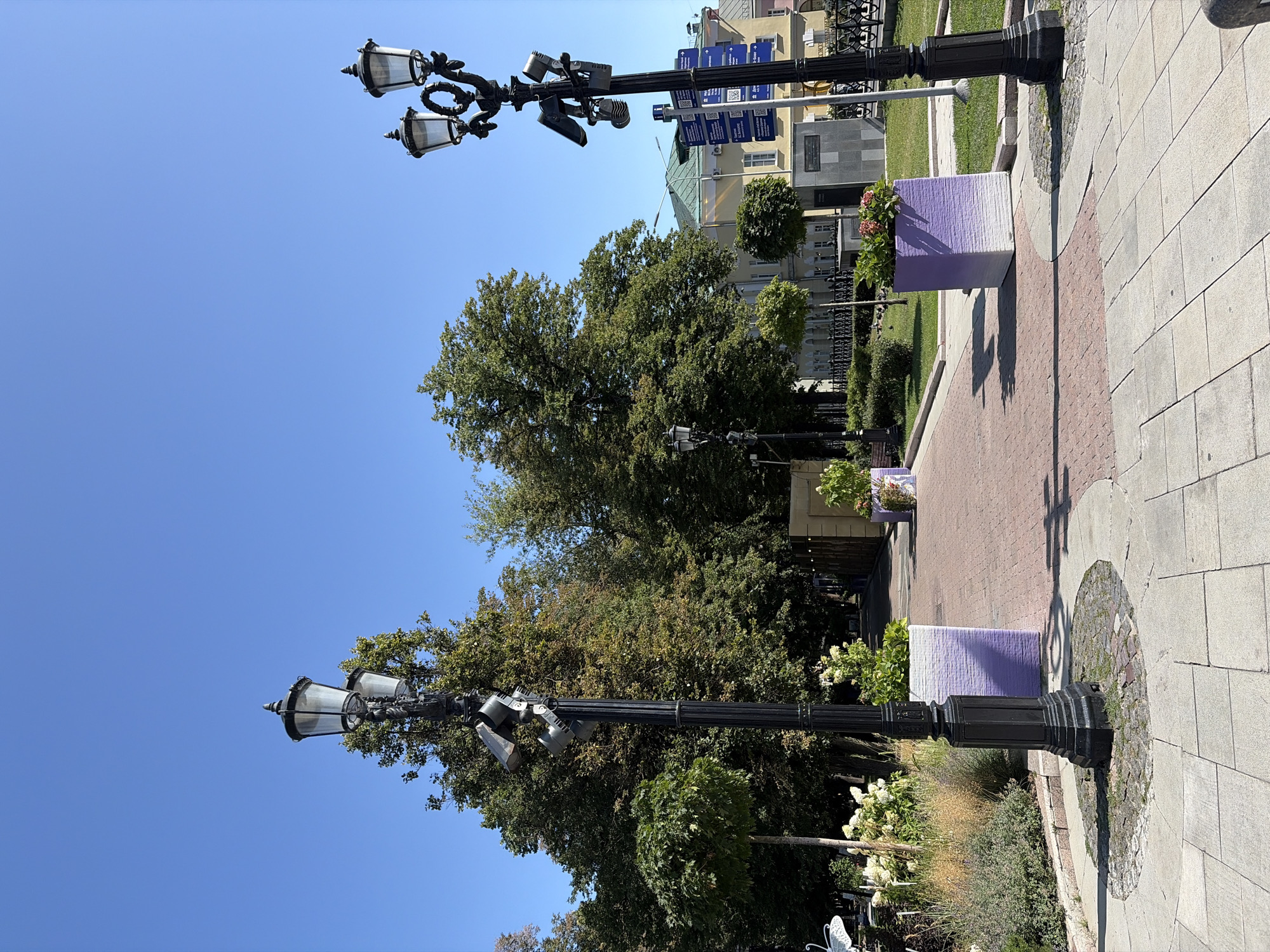 The image captures a sunny day in Moscow, Russia, specifically within a park or pedestrian walkway. Two ornate black lampposts stand prominently in the foreground, each adorned with multiple lamps and additional fixtures, including what appear to be security cameras. The lamppost on the right also features several blue signs with white text and QR codes.
In front of the lampposts are large, rectangular planters with a gradient design transitioning from white to purple, filled with greenery and pink flowers. The walkway is paved with reddish-brown bricks, bordered by lighter-colored flagstones. Lush trees with dense green foliage line the path, creating a dappled shade effect. In the background, a light-yellow building with a green roof is visible, suggesting the proximity of urban infrastructure. The sky is a clear, bright blue, indicative of fair weather. No people are clearly visible in the frame, but a few birds are scattered on the grass. The overall scene depicts a well-maintained public space designed for pedestrian enjoyment and possibly security.