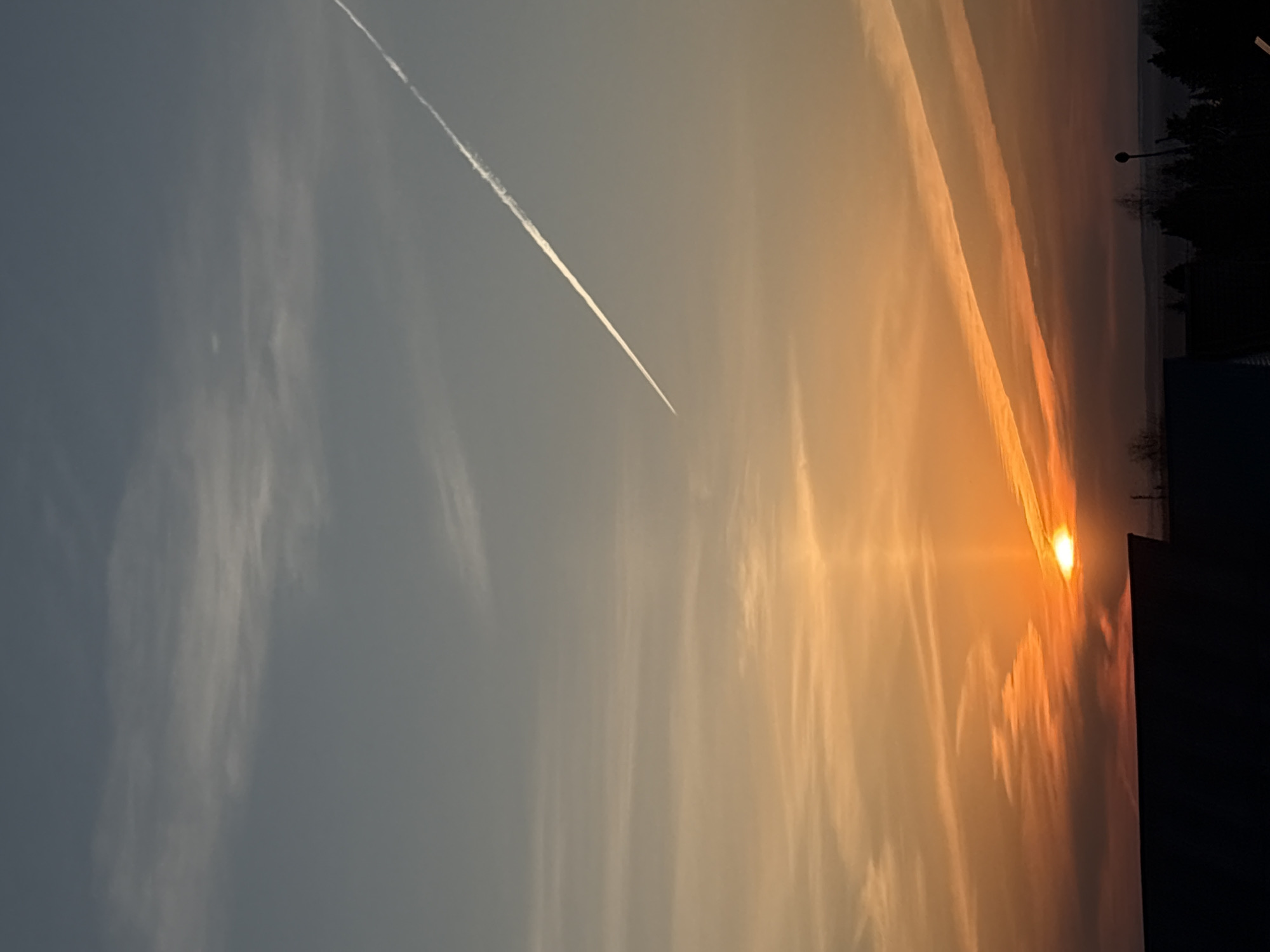 The image depicts a sunset sky in Прикамский, Russia. The sun is near the horizon, creating a bright orange glow, with rays of light streaking upwards. There are high-altitude clouds, some appearing as long, thin streaks reflecting the sunlight. A contrail from an aircraft is visible diagonally across the upper left portion of the sky. In the lower right of the image, silhouetted trees and buildings are discernible against the bright sunset.