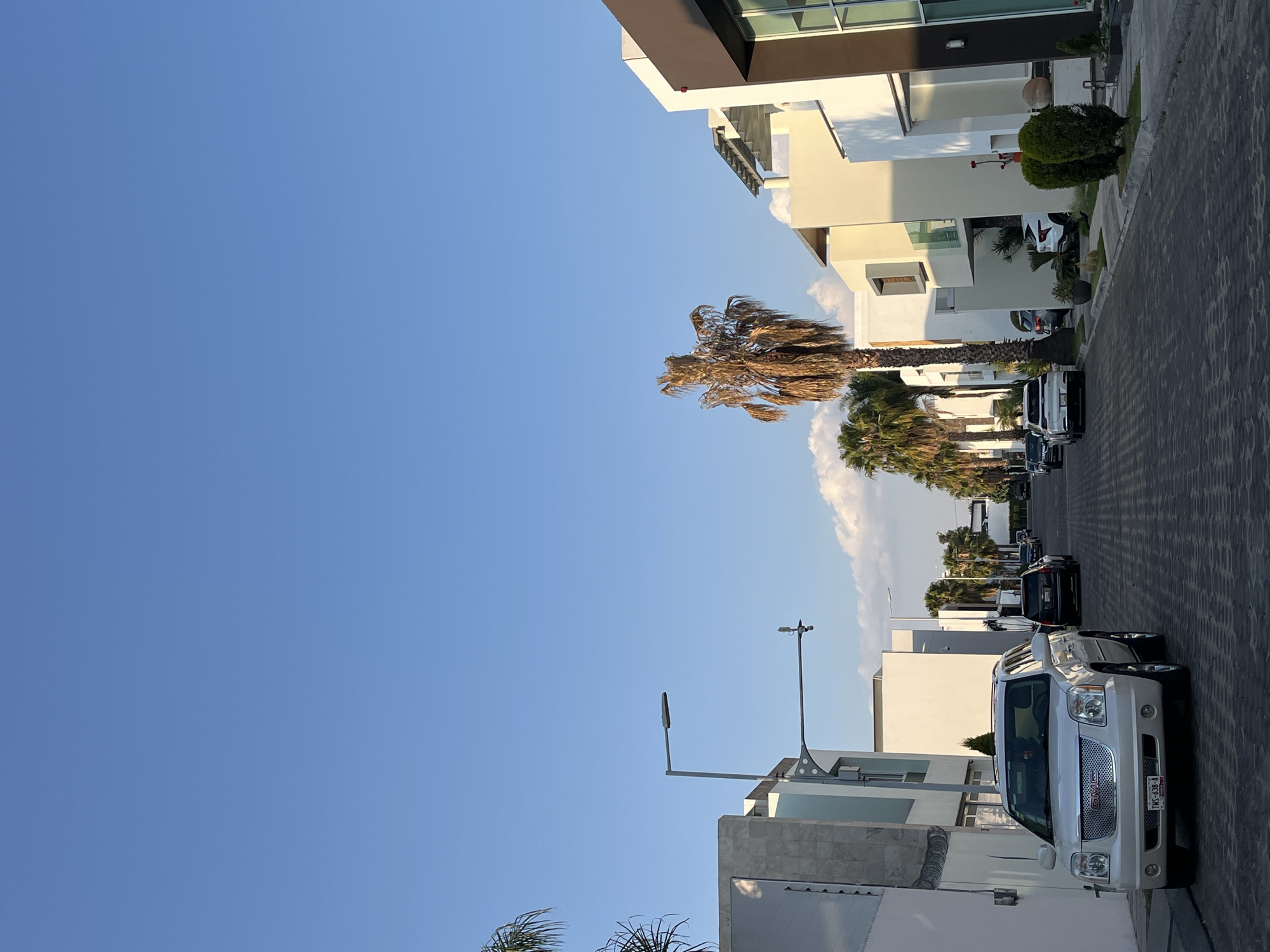 The image shows a street scene in San Andrés Cholula, Mexico, with modern, two-story houses lining both sides. A line of parked cars is visible along the right side of the street, with a white SUV parked prominently in the foreground. Palm trees are interspersed amongst the houses. The sky is a clear, bright blue, suggesting a sunny day. The overall impression is a well-maintained, upscale residential neighborhood.