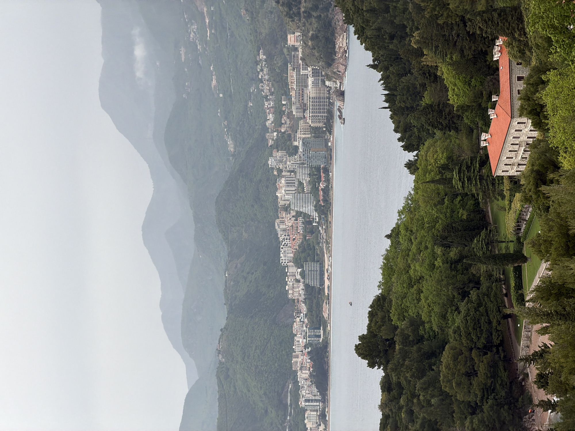 This image, when viewed in landscape orientation, captures a panoramic coastal scene identified as Sveti Stefan, Montenegro, under soft, diffused lighting. On the far left, a series of majestic mountains recedes into the distance, partially obscured by atmospheric haze or mist, creating a layered and somewhat subdued backdrop.
The middle ground features a tranquil stretch of the Adriatic Sea, its surface calm and reflecting the soft light. Along the near shoreline, a significant urban development unfolds, consisting of numerous buildings, likely hotels and resorts. These structures, predominantly white and grey, are densely built along the coast, showcasing a blend of modern architecture against the natural landscape. A small, indistinct boat is visible in the distant water.
To the right, the scene is dominated by a lush, heavily forested hillside that extends towards the sea. Nestled within this dense greenery, a large, prominent building stands out with its distinctive red-tiled roof, stone-colored facade, and several white-trimmed dormer windows. This structure, likely Villa Miločer, is surrounded by manicured lawns and pathways that hint at an elegant estate or resort setting.
The time of day appears to be daytime, but the overcast or hazy weather conditions lend a muted and serene quality to the entire landscape. There are no individuals or specific activities discernible in the image, emphasizing the grand scale and natural beauty of the location. No visible text is present.