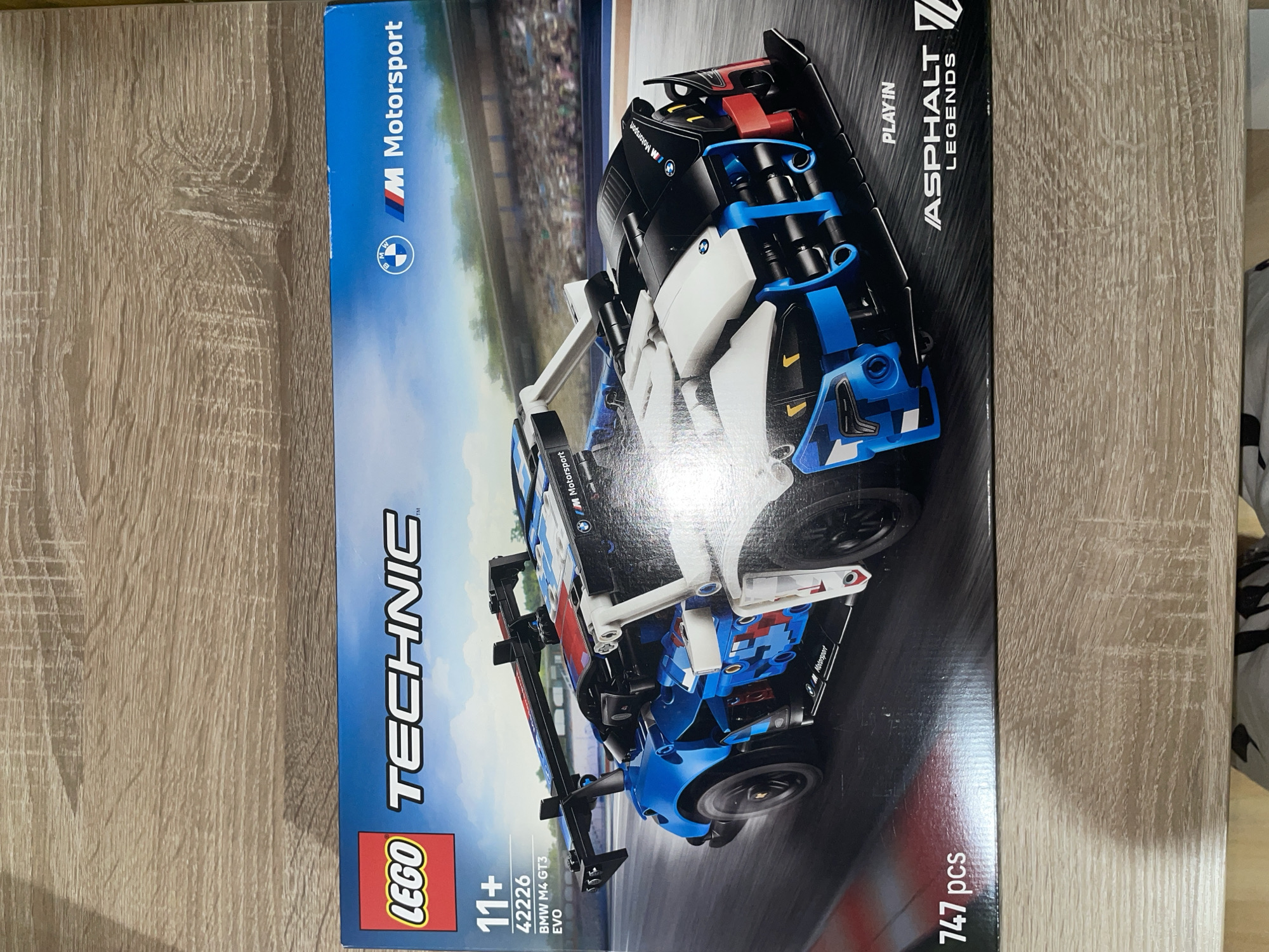 The image shows a LEGO Technic set of the BMW M4 GT3 EVO. The box features the Lego logo, the Technic logo, the age recommendation (11+), the set number (42226), the car model (BMW M4 GT3 EVO), the piece count (747 pcs), and the logos for "M Motorsport" and "Asphalt Legends". The background of the box depicts a racetrack. The box is placed on a wooden surface. The image does not contain information about the location being in Sofia, Bulgaria, nor any information about any people or specific interactions, so I am unable to include those details.