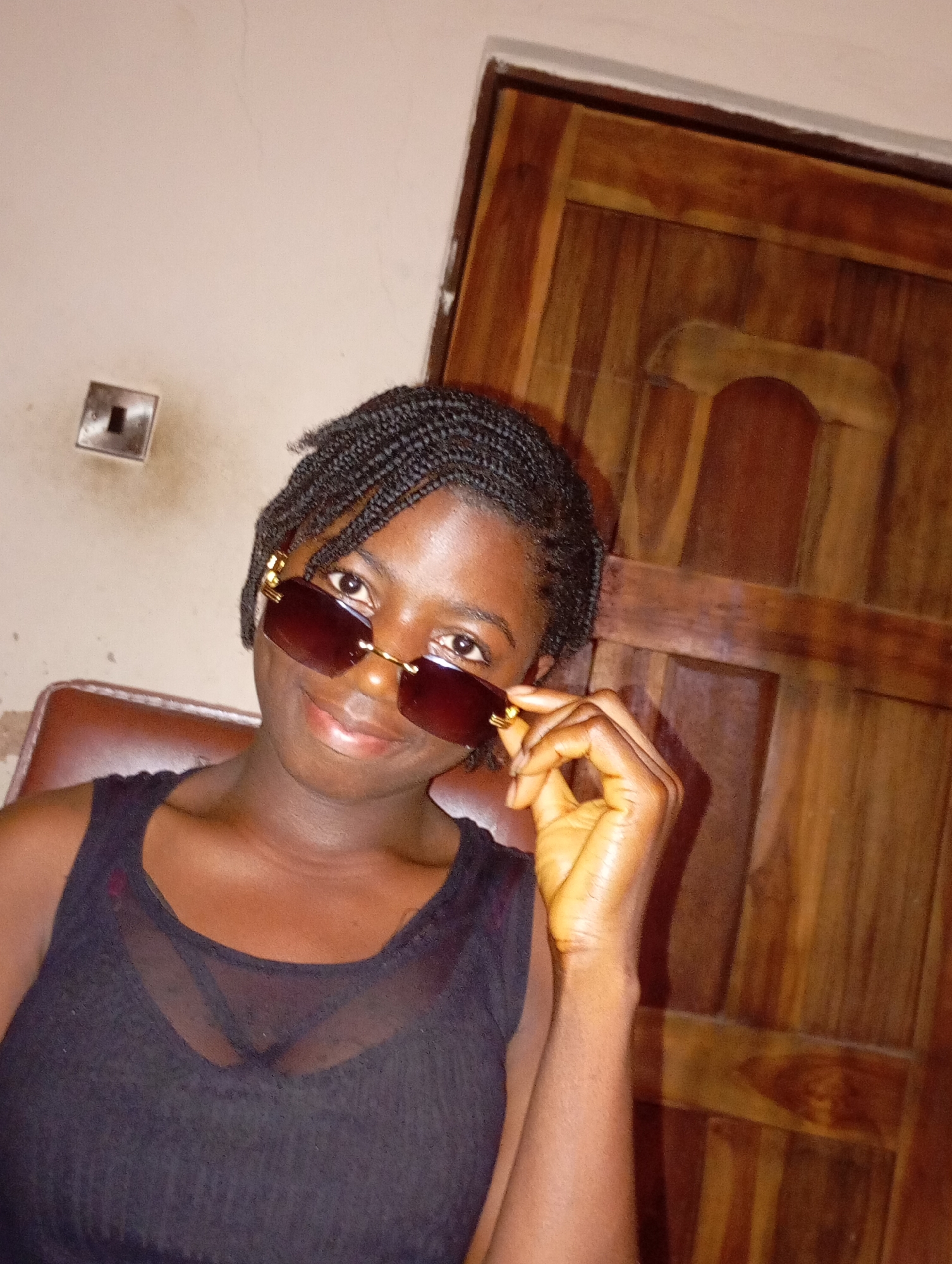 A woman with braided hair is wearing sunglasses and a black tank top. She is sitting in a chair, with a wooden door and a light switch in the background. The location is Unknown, Unknown.