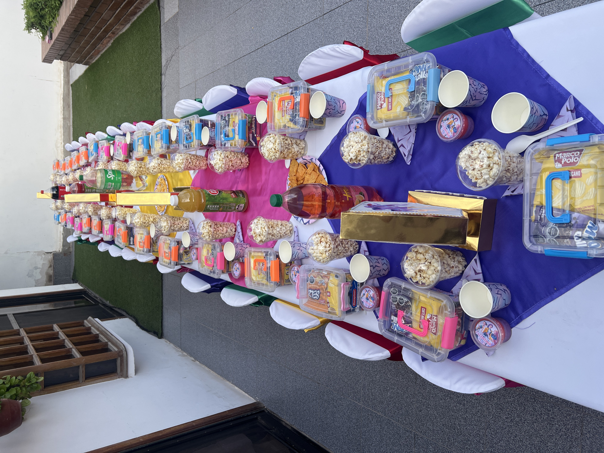 Here is a detailed description of the image:
The image shows a long table laden with party favors and snacks, set outdoors on a patio. The table is covered with a colorful tablecloth that features sections in blue, pink, yellow, and white, with decorative trim around the edges.
Along the length of the table, there are numerous individual snack setups, each apparently intended for one person. Each setup includes items like:
* A clear plastic container filled with chips of various brands.
* A paper cup, some of which are decorated.
* A small container with a character logo, likely holding a treat or toy.
* A plastic cup filled with popcorn.
* Juice bottles are placed intermittently along the table.
* Golden boxes are placed intermittently along the table.
The overall impression is of a party setting, possibly a birthday celebration or similar event, with individual portions of food and treats prepared for attendees. The table is set on a patio with gray paving stones. A building is visible on the left, and artificial turf is visible in the upper left corner. The photo seems to have been taken in the city of Arica, Chile, based on textual references (like "Marco Polo" and brand logos) present on the items.