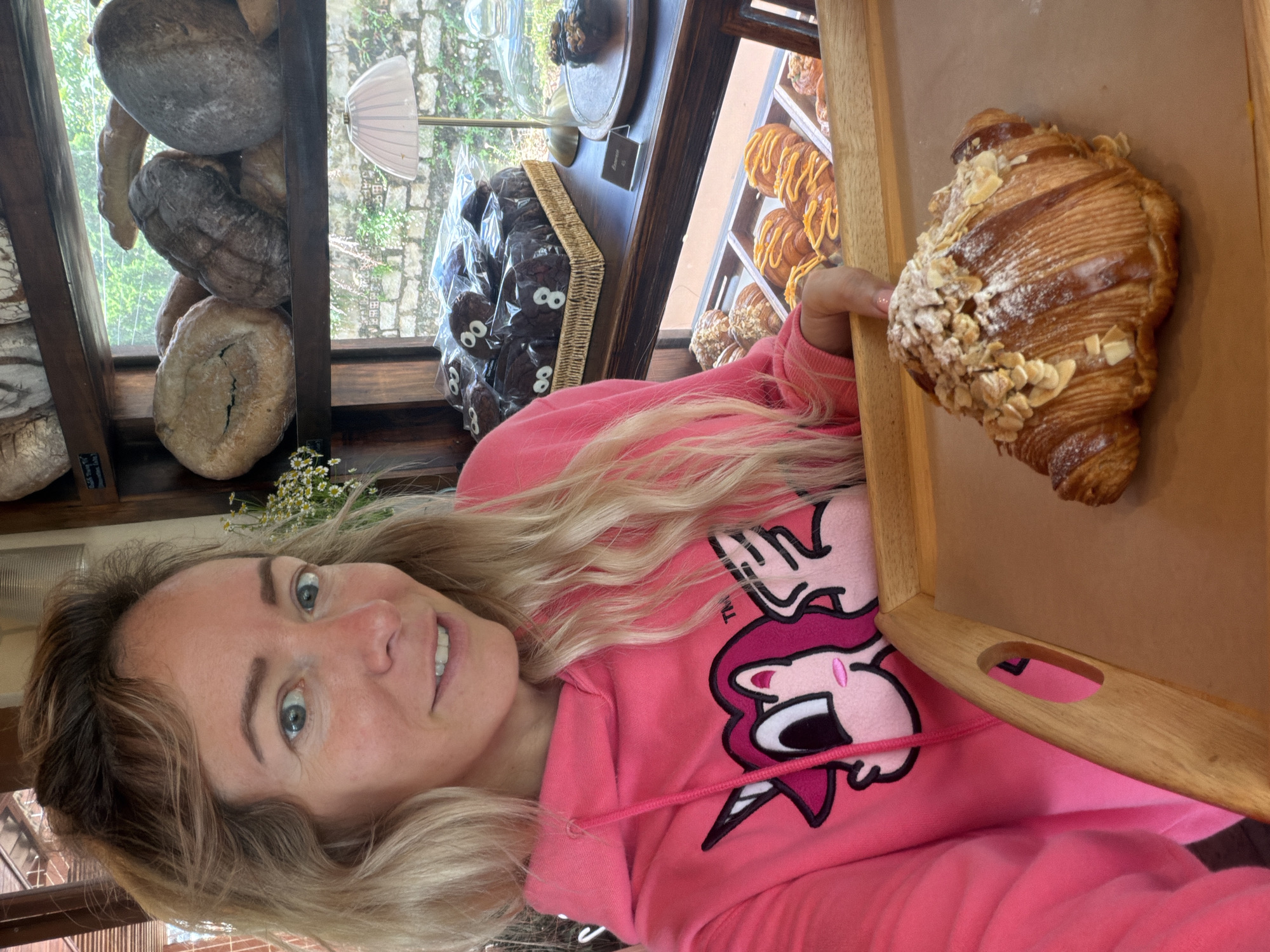 A woman with blonde hair and blue eyes, wearing a bright pink hoodie with a unicorn graphic, is taking a selfie in a bakery. She is holding a wooden tray with a croissant dusted with powdered sugar and almond flakes. Behind her, shelves are stocked with various loaves of bread and pastries. In the background, a window shows a glimpse of greenery and a brick wall, characteristic of the architecture found in Da Lat, Vietnam. The overall scene suggests a casual, friendly atmosphere in a local bakery.