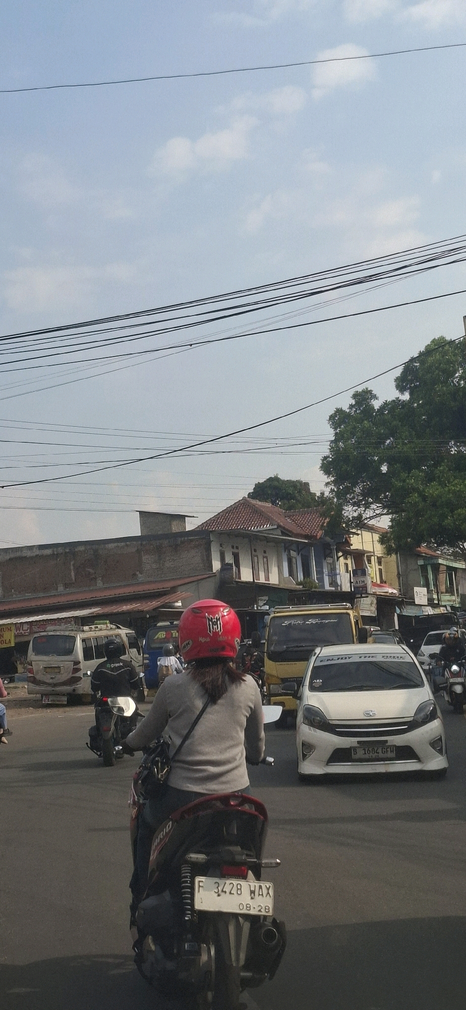 In Sirnagalih, Indonesia, a person wearing a red helmet rides a motorcycle in traffic. The rider is facing away from the camera, wearing a gray long-sleeved shirt and blue jeans, with a black bag slung across their shoulder. The motorcycle's license plate reads "F 3428 WAX" and "08-28". Other vehicles are visible on the road, including a white car with the license plate "B 1602 GFW" and a yellow truck. Buildings line the street, with power lines overhead against a bright, partly cloudy sky. The scene depicts a typical day in a bustling Indonesian town.