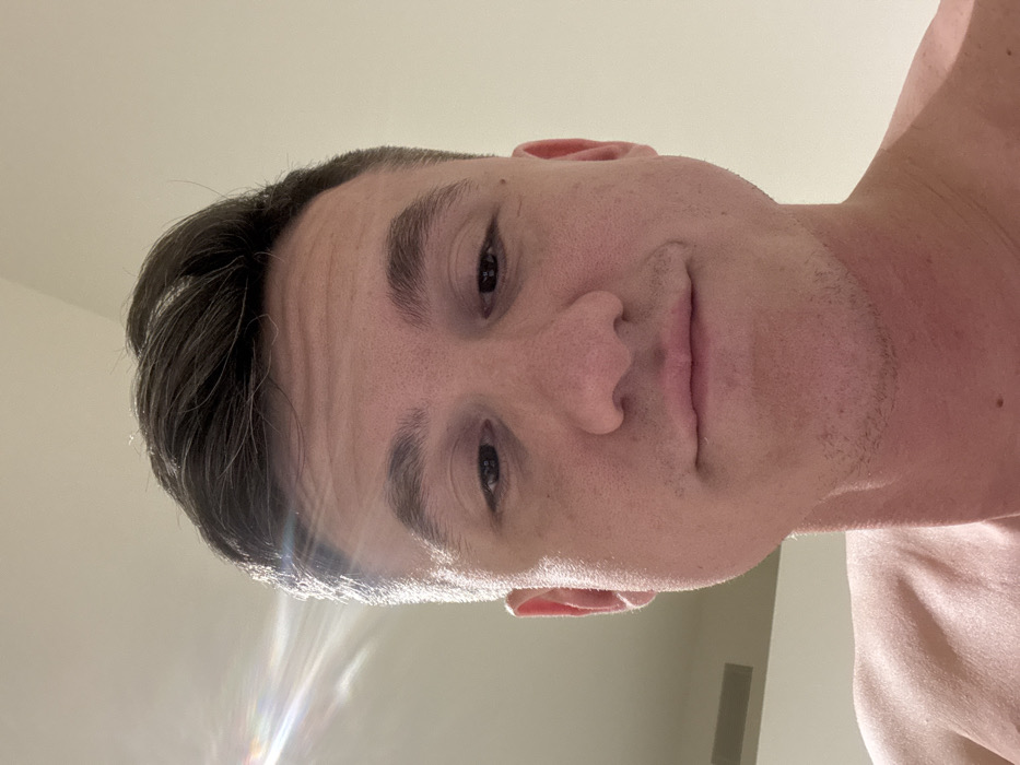 The image displays a close-up, upward-angled portrait of an adult male. The subject has short, dark hair, dark eyes, and light stubble on his lower face. He is looking directly at the camera with a neutral expression, exhibiting a subtle curve at the corners of his mouth. A portion of his bare shoulder is visible in the lower-left quadrant, indicating he is shirtless. The background consists of a plain, light-colored interior wall or ceiling. A prominent lens flare from an unidentified light source is visible in the upper-left corner of the frame. A rectangular ventilation grille is partially visible in the lower-left background, suggesting an indoor environment. No visual cues within the image definitively place the location in the city of Venice, Italy.