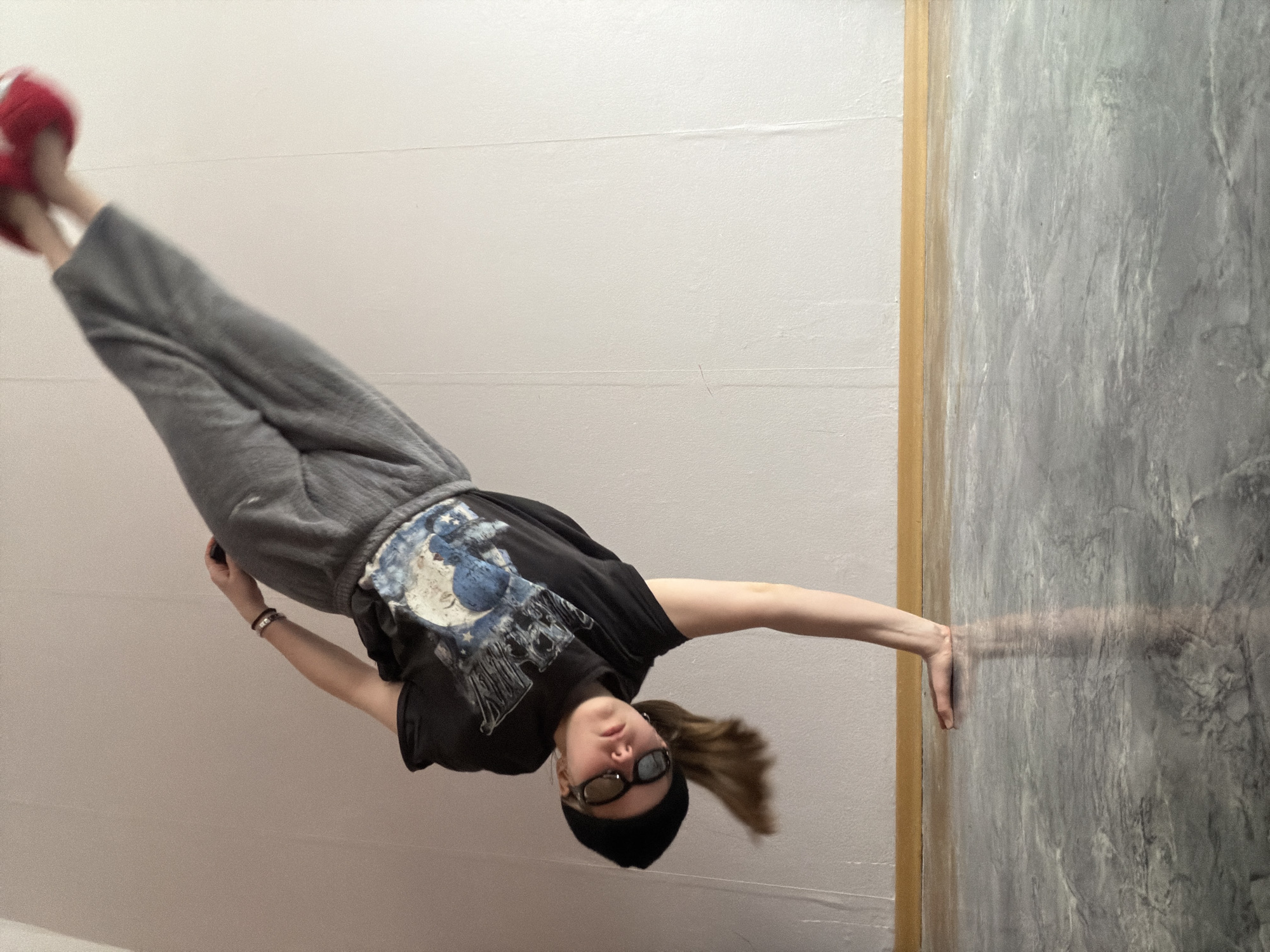 The image captures a person, likely female, in a dynamic and unusual pose indoors in Perm, Russia, creating the illusion of floating or defying gravity. The subject is positioned diagonally across the frame, leaning into the scene.
She is dressed casually in a black t-shirt featuring a blue and white graphic design, grey sweatpants, and distinctive red shoes or slippers. Her head is adorned with a black beanie, and dark sunglasses obscure her eyes, giving her a cool, enigmatic appearance. Her light brown hair is visible behind her head, exhibiting a slight blur consistent with movement or a long exposure. A wristband or bracelet is worn on her left wrist, and she holds a small, dark object, possibly a smartphone, in her left hand.
Her right arm is fully extended, with her open hand pressed flat against a textured, dark grey wall on the right side of the frame. This interaction with the wall appears to be key to the suspended illusion. Adjacent to this, on the left, is a plain, light off-white or pale grey wall. A thin, vertical strip of light brown material, possibly wood, acts as a divider between the two distinct wall surfaces. Her legs are bent at the knees, with her red-shoed feet pointing upwards towards the corner of the frame. The floor is visible at the bottom edge, also light-colored.
The image captures a moment of playful activity, utilizing perspective to create an artistic or acrobatic impression. No explicit text is visible on the person or in the setting. The indoor lighting provides no specific cues about the time of day.