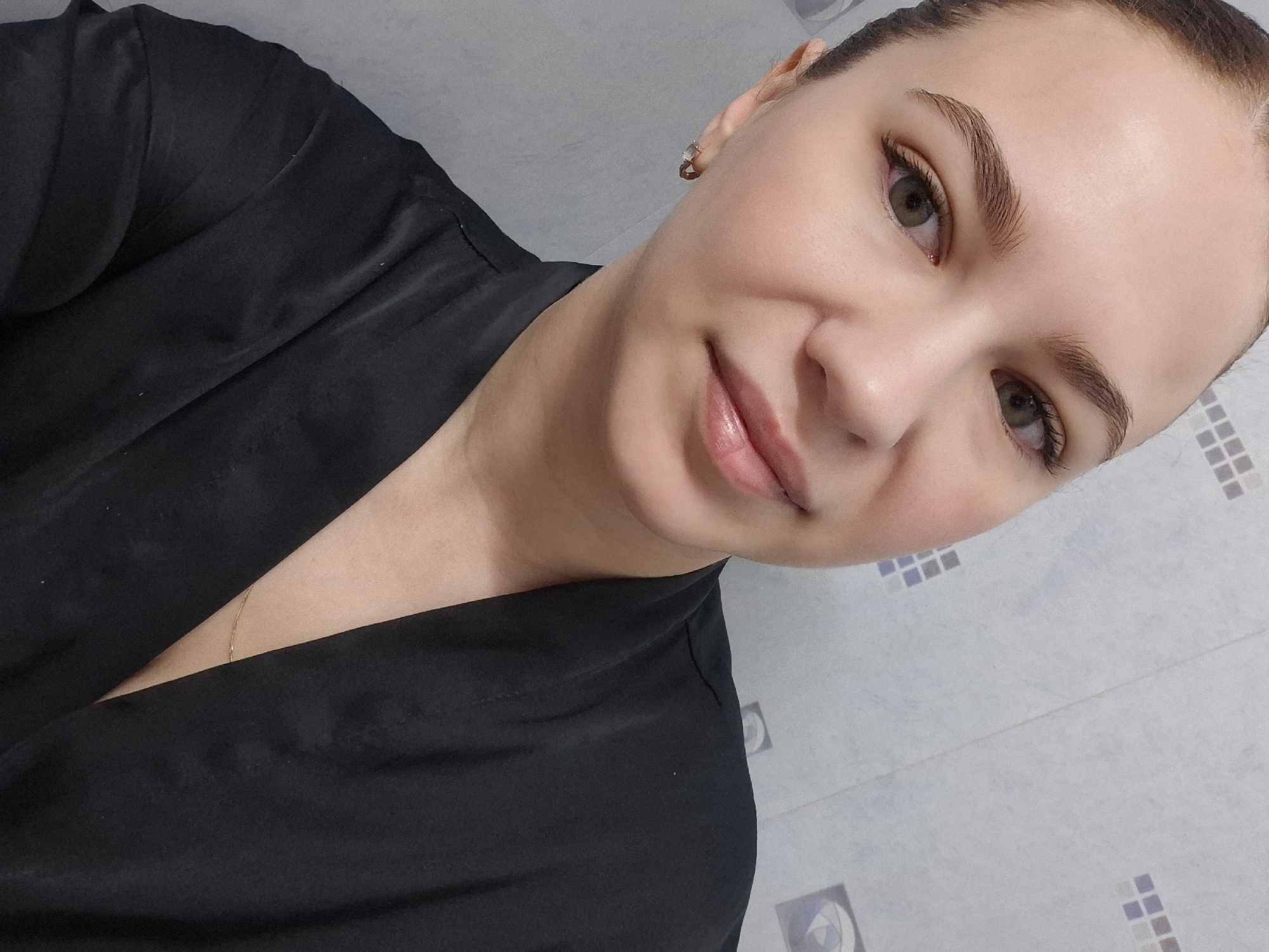 A woman with fair skin, brown hair pulled back, and light brown eyes is looking at the camera. She is wearing a black garment that drapes open to reveal a glimpse of a thin gold necklace. She has subtle makeup on, including defined eyebrows and glossy lips. She is wearing gold stud earrings. The background consists of a wall with a patterned, tiled surface in shades of white and blue. The location is identifiable as the city of Chaikovsky, Russia, but specific details of the location within the city are not discernible from the image.