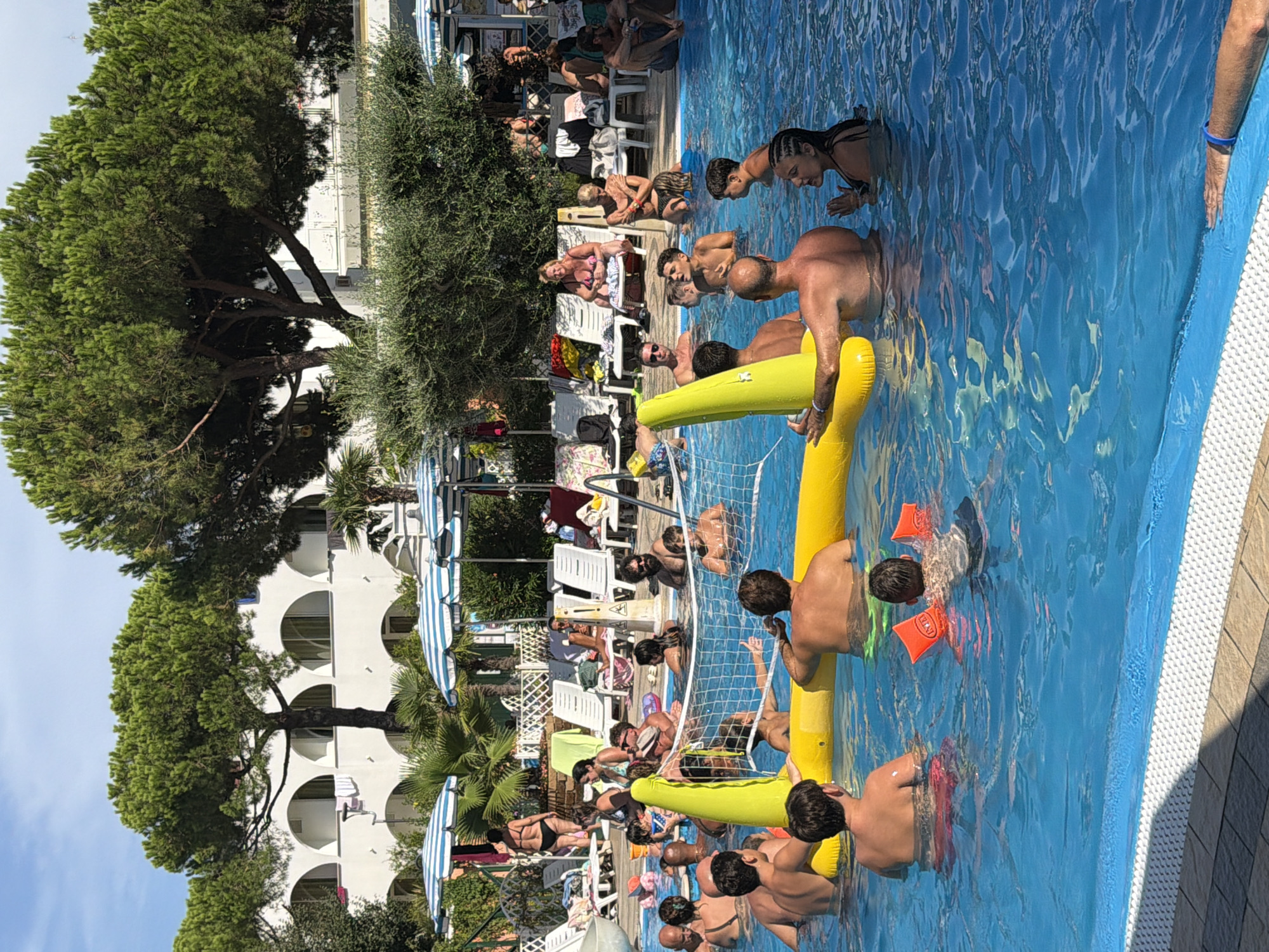 In the city of Gatteo, Italy, a vibrant scene unfolds at a poolside, where people are enjoying a sunny day. The azure water of the swimming pool is populated with individuals of various ages, engaged in leisure activities. In the foreground, a bright yellow inflatable volleyball net stands as a central feature, with several people actively participating in a game. Some are in the water, some are holding onto the net, and others are looking towards the game.
Surrounding the pool, lounge chairs are set up, some occupied by people relaxing and observing the activities. The backdrop features lush green trees and a white building with arched windows, suggesting a resort or hotel setting. The atmosphere is one of joyful recreation and relaxation, with the clear blue sky overhead indicating pleasant weather. The image captures a moment of communal enjoyment in a warm, Italian setting.