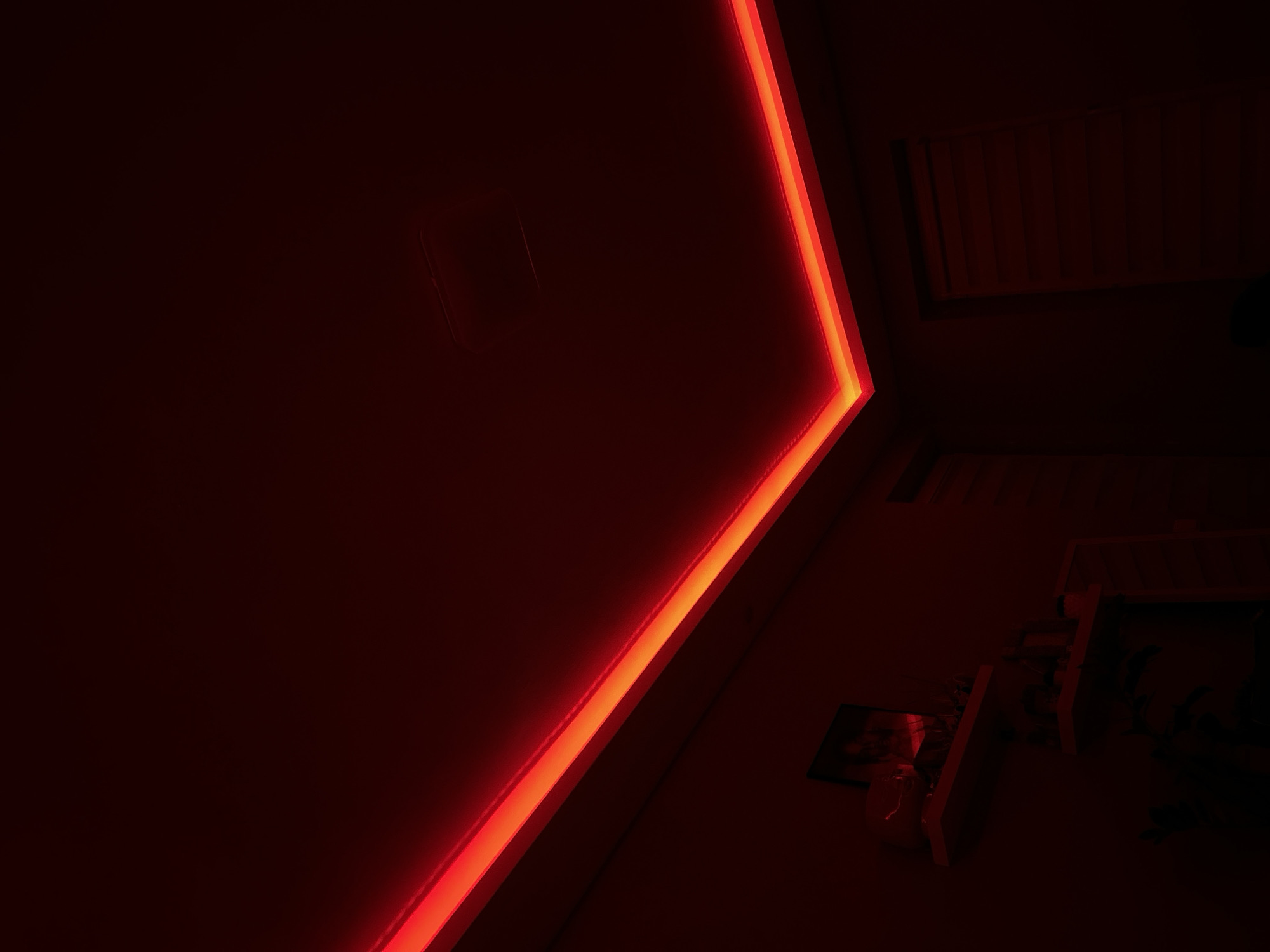 The image depicts an interior space illuminated primarily by red light. A bright red linear light fixture is prominently featured, tracing the corner of a wall. A rectangular indentation, possibly a control panel or access point, is visible on the wall. The environment is darkly lit, with some furniture or equipment visible in the background, including what appears to be a shelf. The scene is too dark to definitively identify the location within Goce Delcev, Bulgaria, without further visual or contextual information.