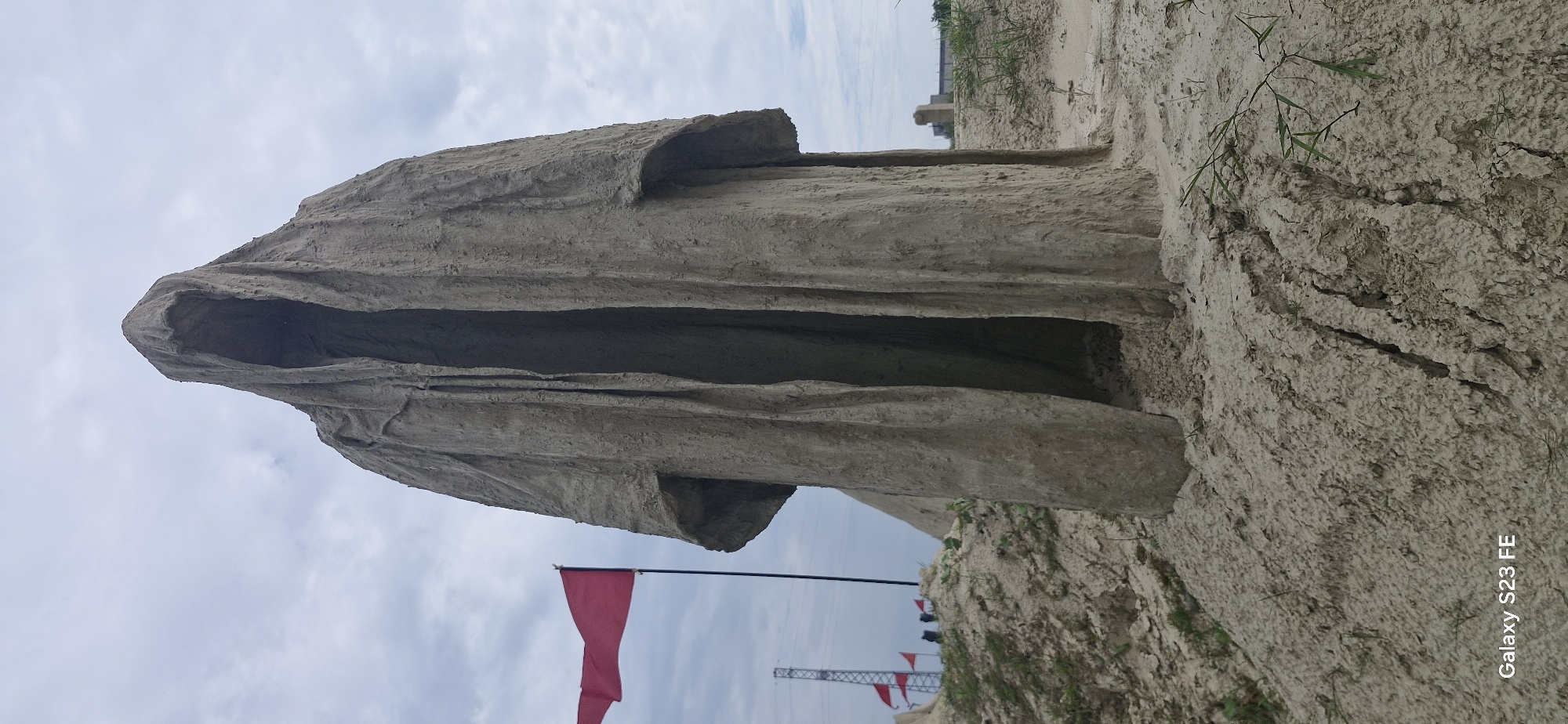 The image shows a large, cloaked statue made of a rough, sandy material, positioned outdoors on a sandy or earthen embankment. The statue has a hooded head with a dark, hollow opening where a face would be, and its body is draped in a heavy, robe-like garment with deep folds. The texture of the statue suggests it is made of clay or a similar malleable substance that has dried and hardened. In the background, a red flag on a pole is visible to the left of the statue, slightly blurred. Further in the background, there are industrial structures, including a tall lattice tower with lights. Other flags and small figures of people are present on a distant hill. The sky is overcast with clouds. The foreground shows the sandy ground with some sparse vegetation. The image is a vertical, full-shot of the statue. The bottom left corner of the image displays "Galaxy S23 FE". The statue is located on the territory of the former Jelgava Prison island (Kalnciema iela 60A, Jelgava, Latvia).